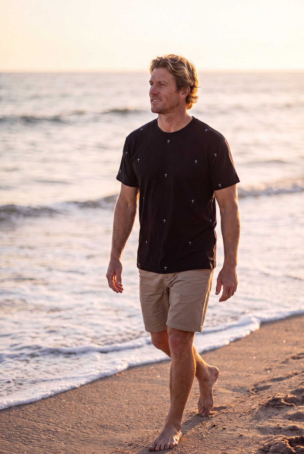 A man walks on a sandy beach at sunset wearing The Cross classic fit ring spun tee in black-hand illustrated cross pattern-Fox and Form. The Men's Signature Collection. 