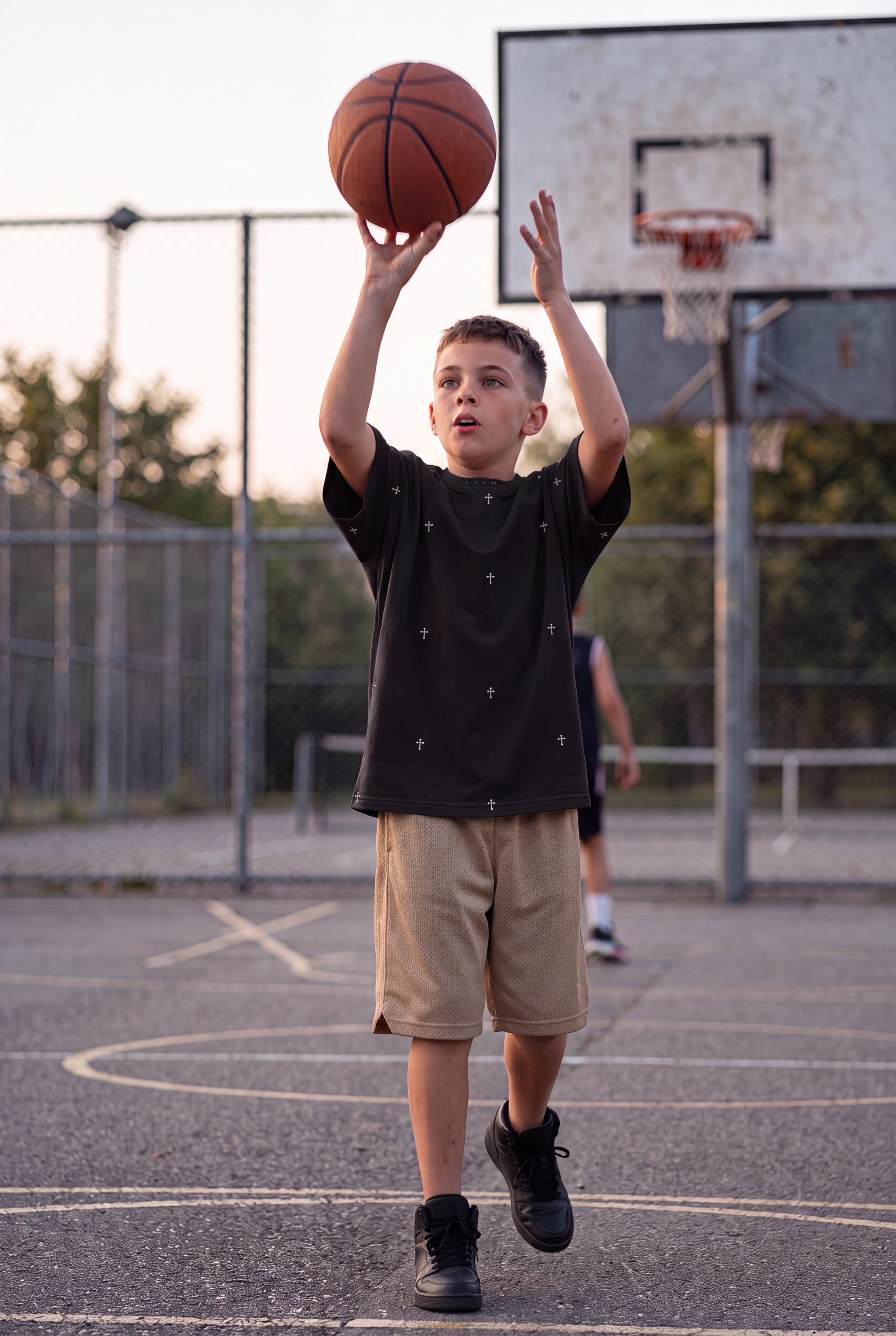 Young boy shooting a basketball at an outdoor court wearing Fox and Form The Cross black dropped shoulder max weight tee with hand-etched illustrated cross pattern design