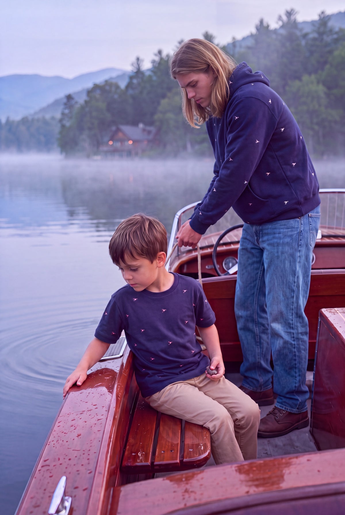 A man and a boy wearing matching navy-blue hoodies with small brown fly-fishing lure patterns are on a wooden boat on a misty lake with mountains in the background. Fox & Form. Mini & Youth Signature Collection.