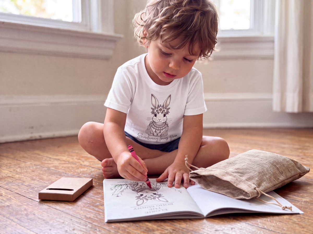 A toddler sits cross-legged on a wooden floor in a bright room, drawing in an open sketchbook with a red pencil. The child is wearing a white graphic T-shirt with a rabbit illustration and text, dark shorts, and a beige canvas drawstring pouch lies beside the book.