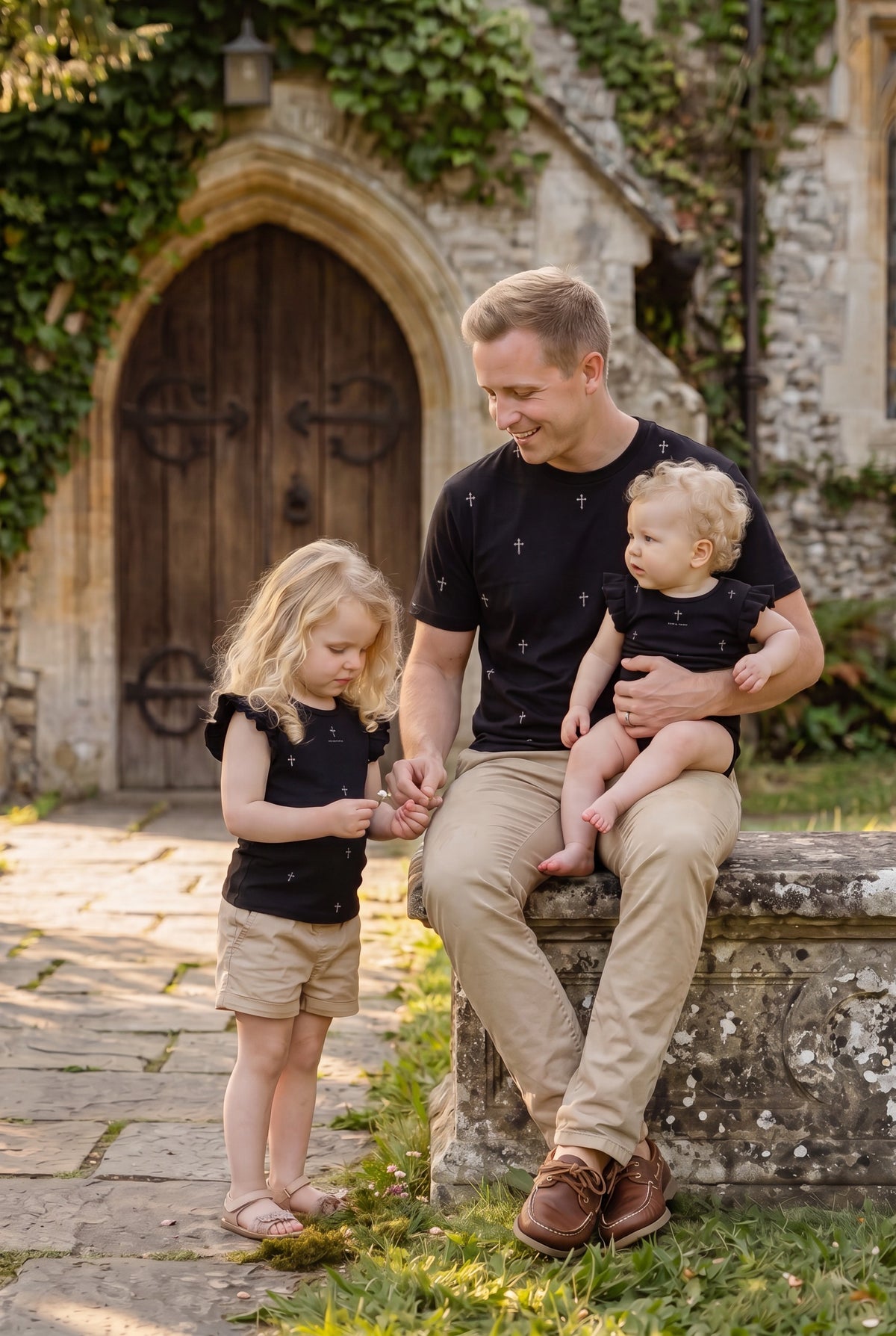 Father holding infant and sitting with toddler daughter outside a stone chapel wearing matching Fox and Form The Cross black classic ring spun tee with hand-etched illustrated cross pattern design. Fox and Form. Mini &amp; Youth Signature Collection. 