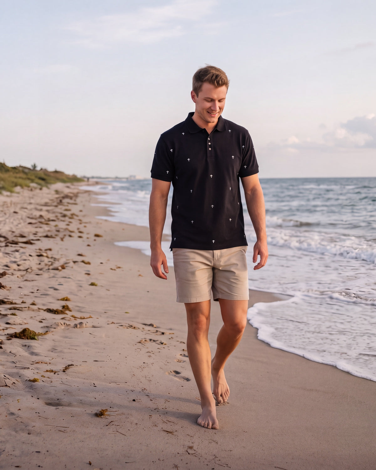 A man walking barefoot on a sandy beach next to the ocean, wearing a black short-sleeve polo shirt with small white, grey, and black hand-etched crosses on front and back and beige shorts. Fox & Form, Signature Collection. 