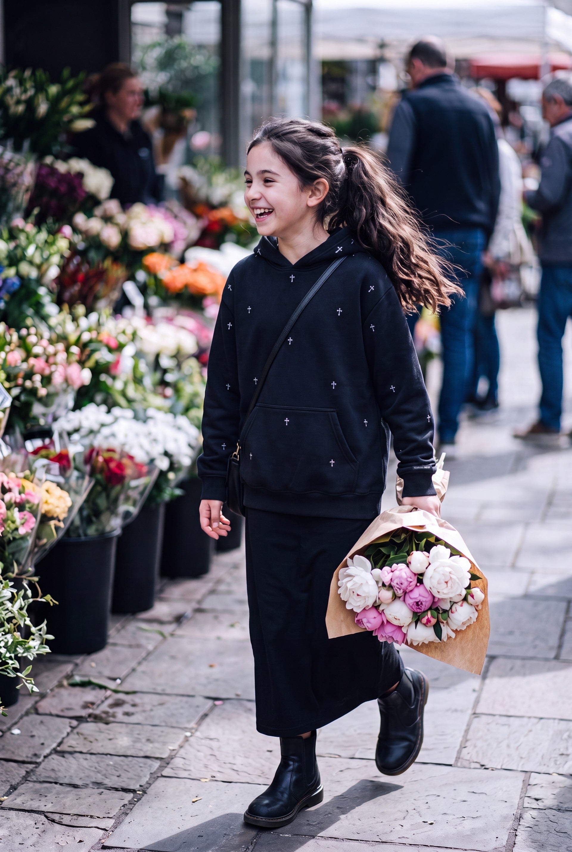 Young girl wearing Fox and Form The Cross black max weight hooded sweatshirt with hand-etched illustrated cross pattern design, browsing at an outdoor flower market. Fox and Form. Mini & Youth Signature Collection. 