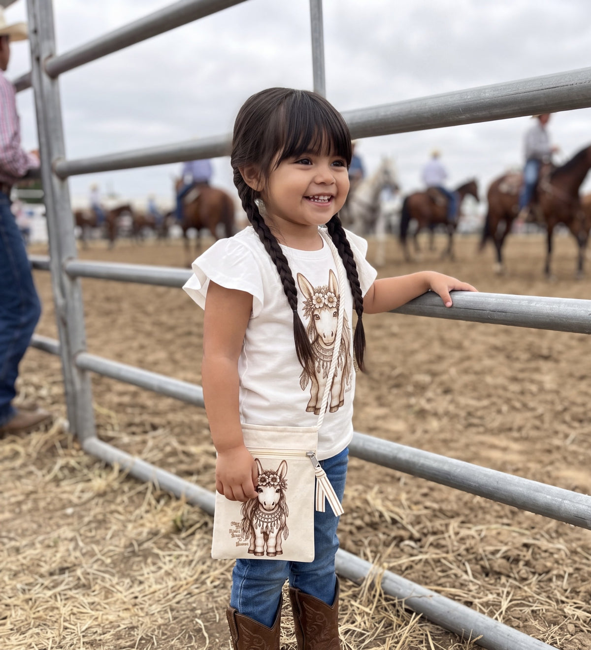 A young girl with braided hair stands in a cowboy arena holding a canvas bag with a brown hand etched meadow the mini boho pony illustration. She is wearing a white t-shirt with a matching hand etched Meadow the mini boho pony illustration graphic, blue jeans, and brown boots, with horses and people blurred in the background behind a metal fence.