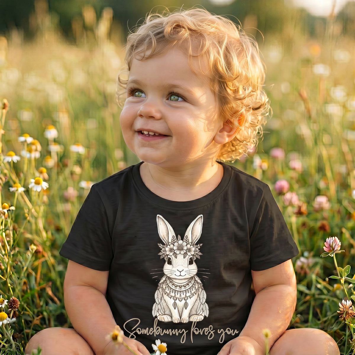 Smiling baby boy sitting in a wildflower meadow at golden hour wearing Fox and Form Elorie the Mini Boho Bunny black classic ring spun tee with hand-etched illustrated boho bunny design and Somebunny Loves You script