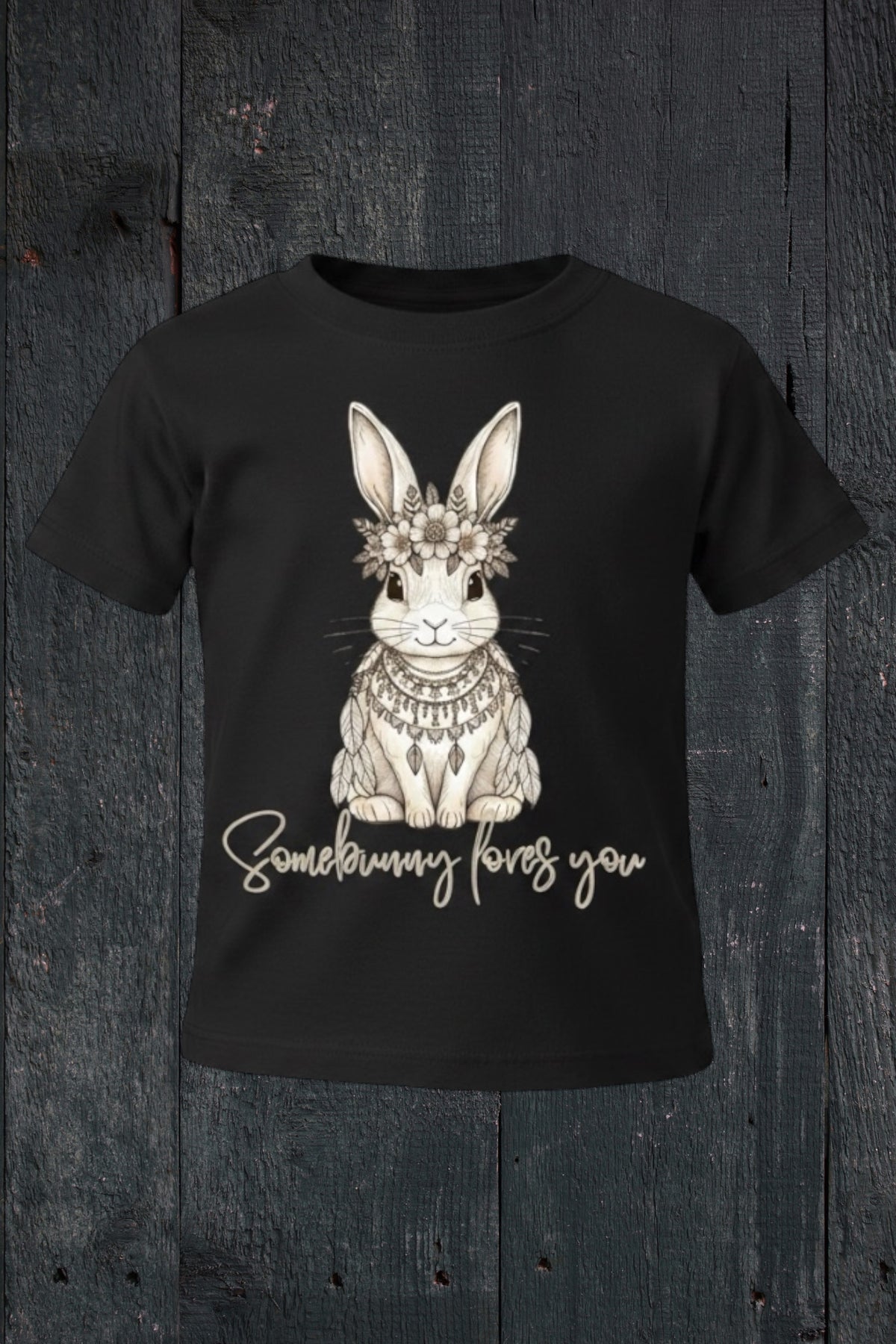 Flat lay of Fox and Form Elorie the Mini Boho Bunny black classic ring spun tee with hand-etched illustrated boho bunny design and Somebunny Loves You script on a dark wood background