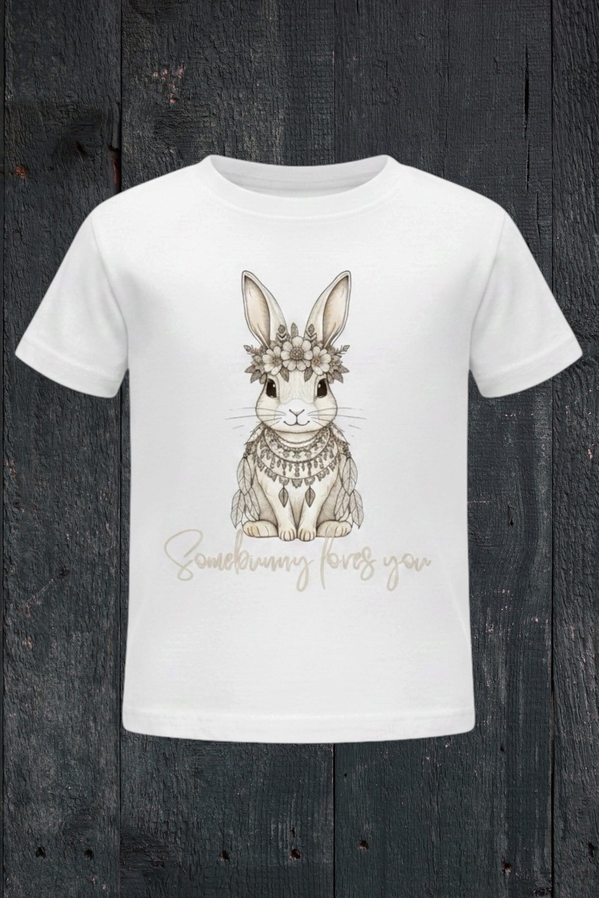 Flat lay of Fox and Form Elorie the Mini Boho Bunny white classic ring spun tee with hand-etched illustrated boho bunny design and Somebunny Loves You script on a dark wood background