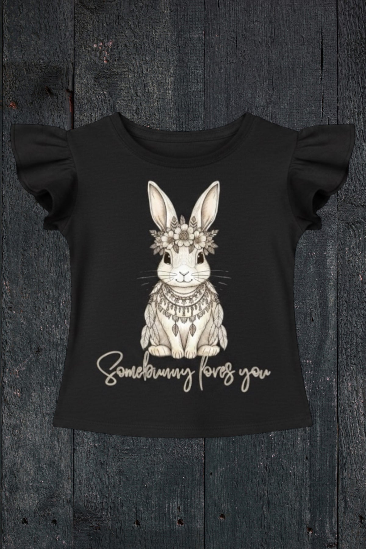 Flat lay of Fox and Form Elorie the Mini Boho Bunny black flutter sleeve tee with hand-etched illustrated boho bunny design and Somebunny Loves You script on a dark wood background