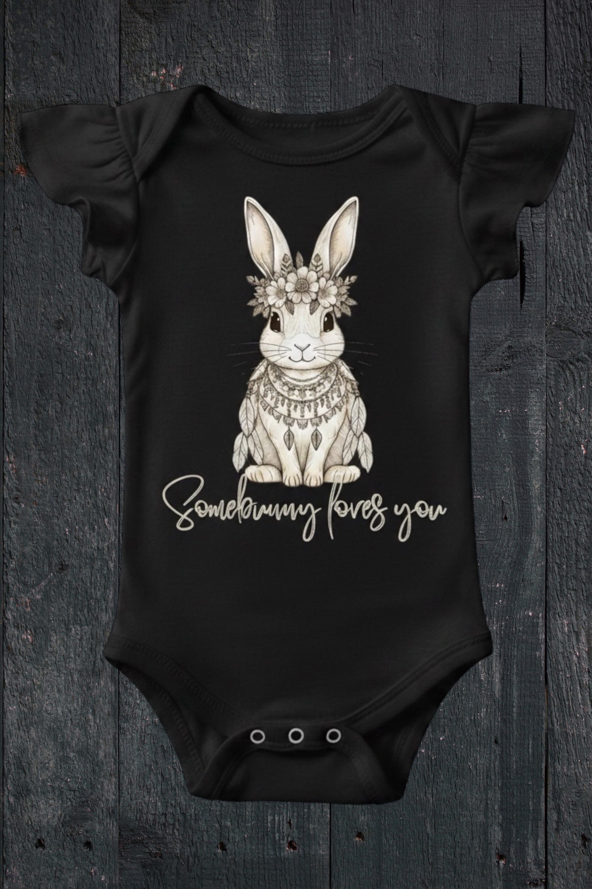 Flat lay of Fox and Form Elorie the Mini Boho Bunny black flutter sleeve onesie with hand-etched illustrated boho bunny design and Somebunny Loves You script on a dark wood background