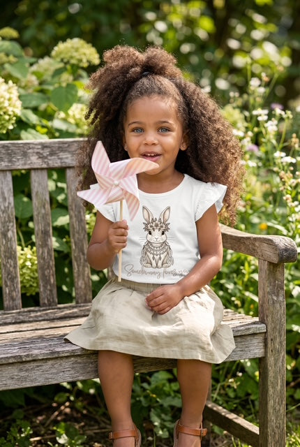Young girl sitting on a rustic wooden garden bench holding a pink pinwheel wearing Fox and Form Elorie the Mini Boho Bunny white flutter sleeve tee with hand-etched illustrated boho bunny design and Somebunny Loves You script