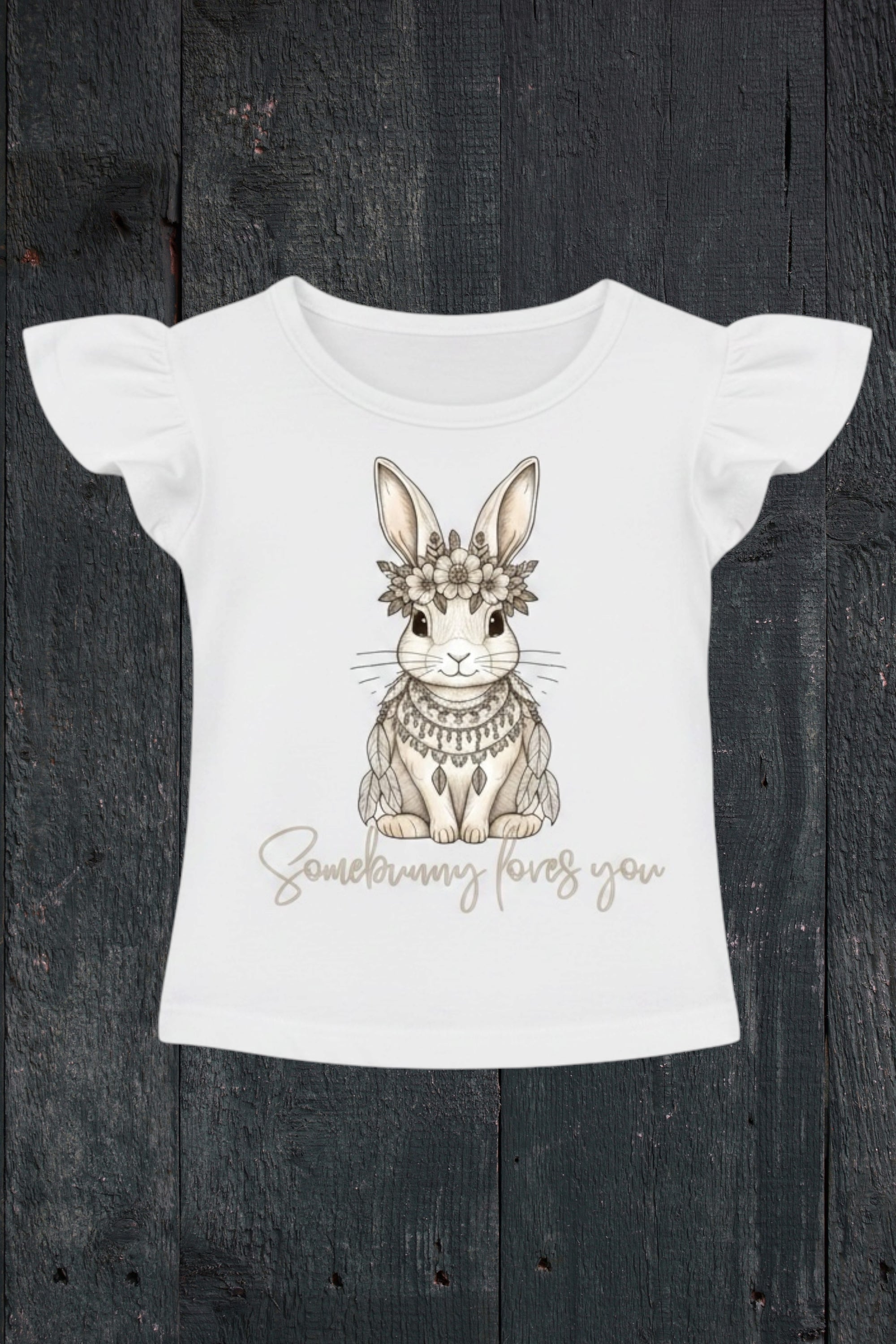 Flat lay of Fox and Form Elorie the Mini Boho Bunny white flutter sleeve tee with hand-etched illustrated boho bunny design and Somebunny Loves You script on a dark wood background