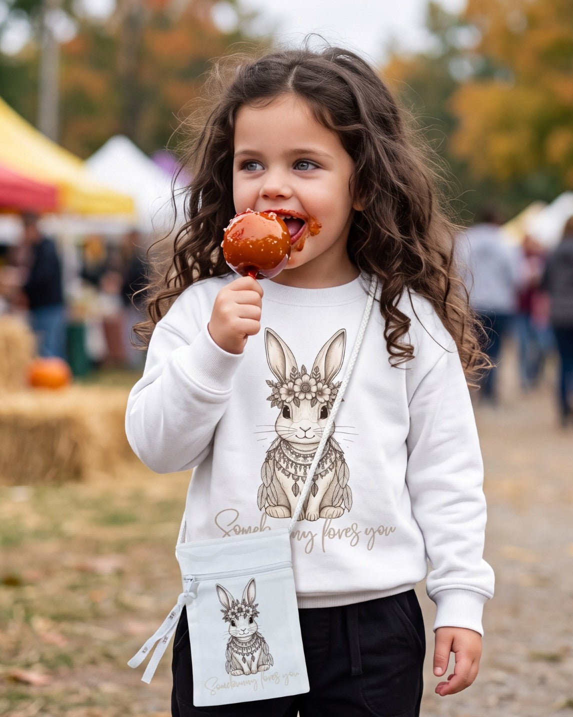 Toddler girl eating a candy apple at an outdoor festival wearing Fox and Form Elorie the Mini Boho Bunny white crewneck sweatshirt with hand-etched illustrated boho bunny design and Somebunny Loves You script and matching crossbody bag. 
