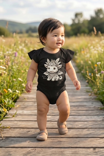 Baby girl taking her first steps on a wooden boardwalk through a wildflower meadow wearing Fox and Form Ember the Mini Boho Dragon black flutter sleeve onesie with hand-illustrated boho dragon design and Born to Soar script