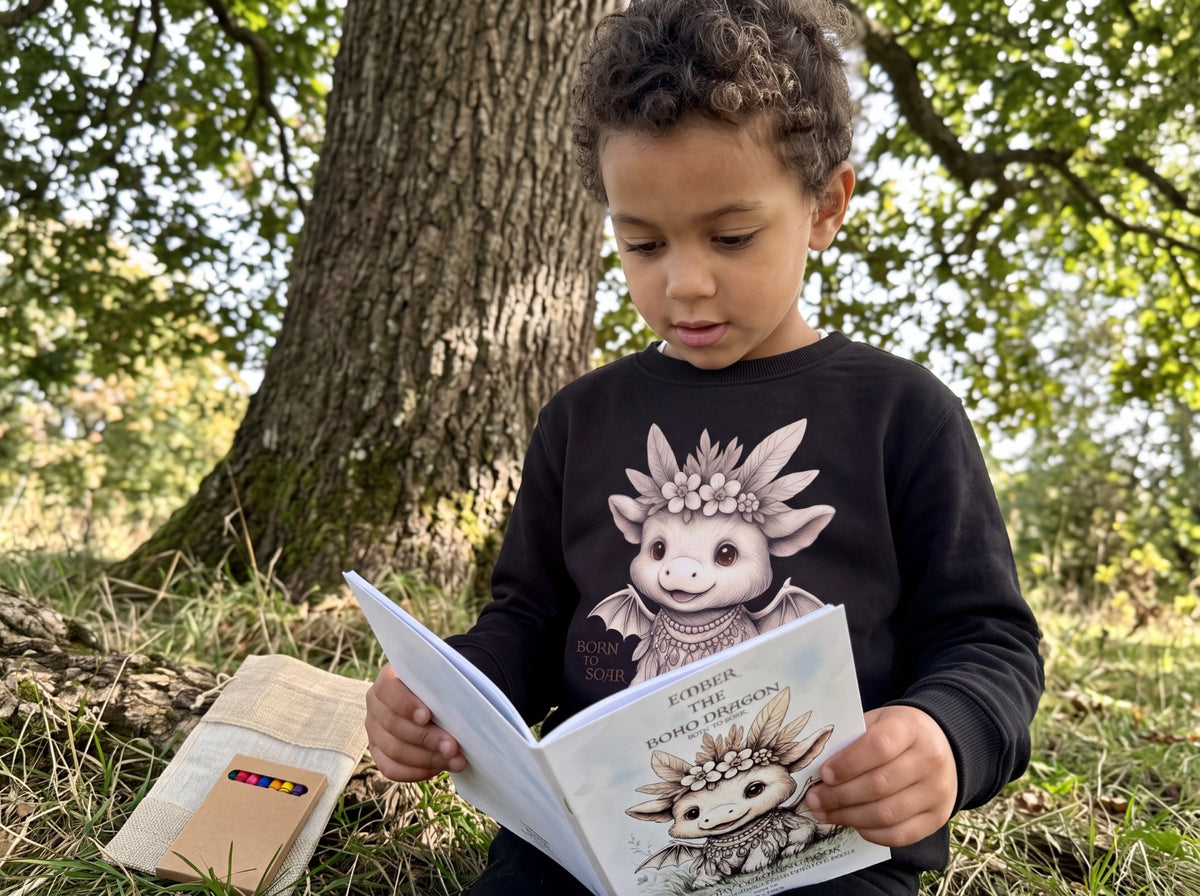 A young child sitting outdoors on the grass in front of a large tree, reading a book about Ember the Boho Dragon by Fox &amp; Form. The child is wearing a black long-sleeve sweatshirt with a detailed illustration Fox and Form Ember the Mini Boho Dragon.