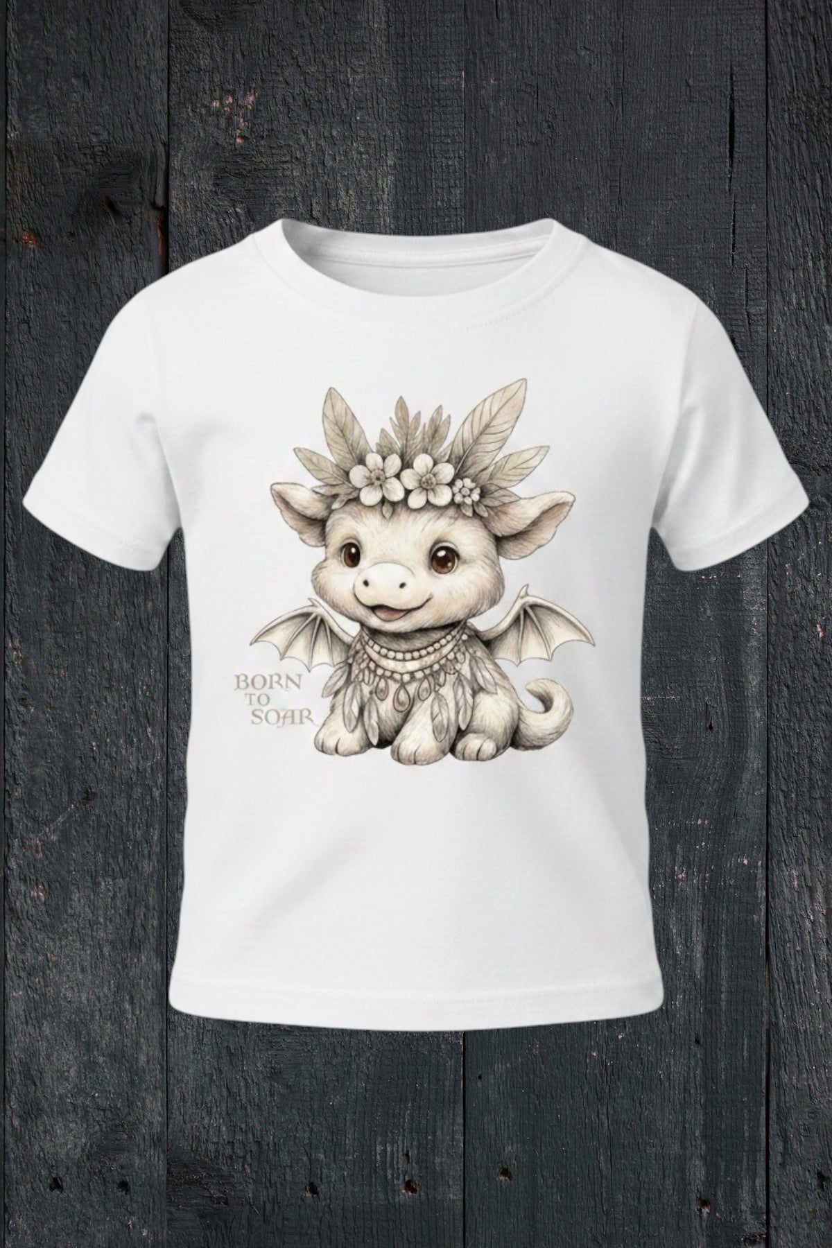 Flat lay of Fox and Form Ember the Mini Boho Dragon white classic ring spun tee with hand-illustrated boho dragon design and Born to Soar script on a dark wood background