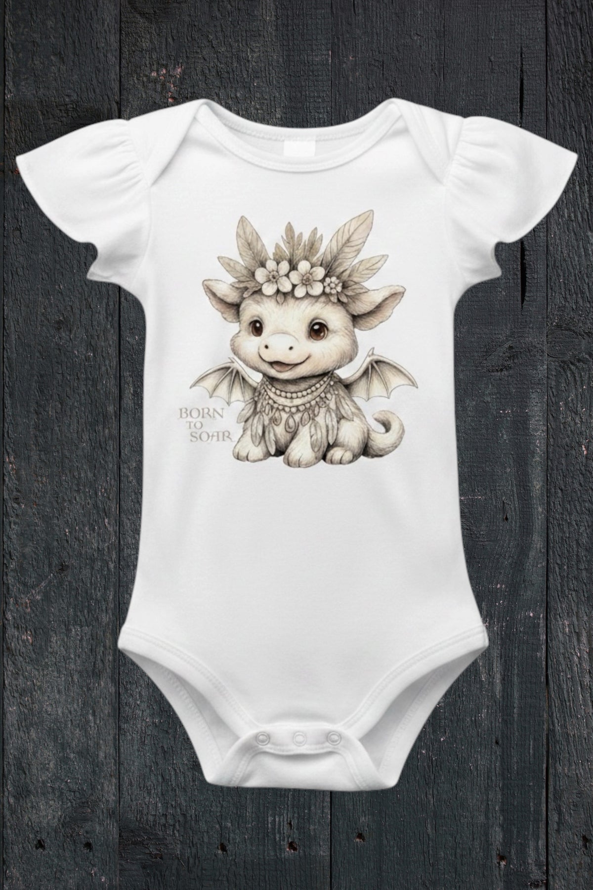 Flat lay of Fox and Form Ember the Mini Boho Dragon white flutter sleeve onesie with hand-illustrated boho dragon design and Born to Soar script on a dark wood background