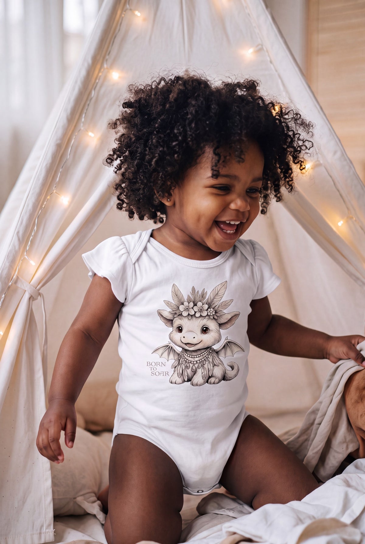 A smiling toddler with curly dark hair wearing a white short-sleeve bodysuit with a cute Ember Mini Boho dragon graphic, sitting inside a white fabric canopy tent with warm string lights. Fox & Form. 