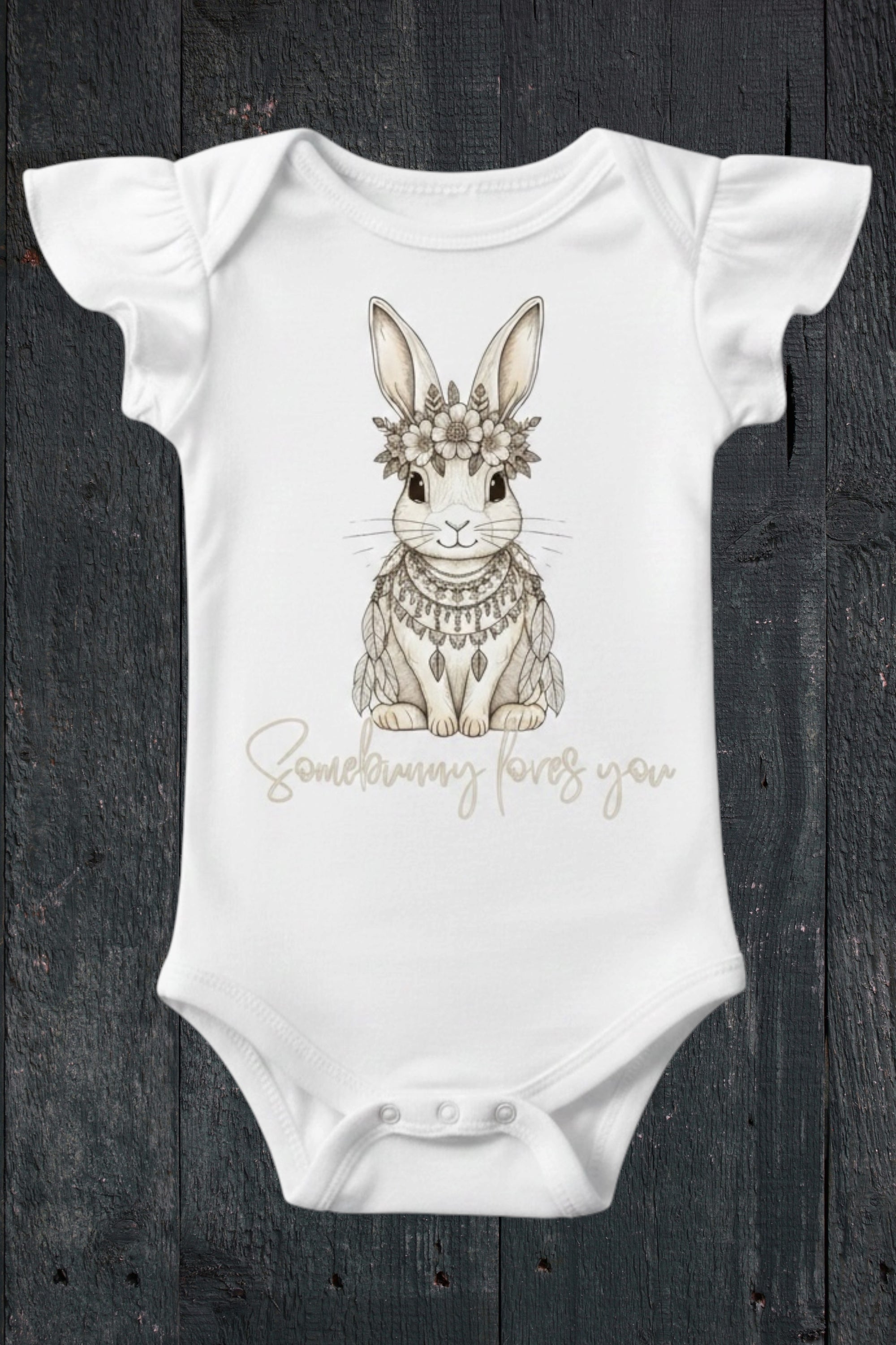 Flat lay of Fox and Form Elorie the Mini Boho Bunny white flutter sleeve onesie with hand-etched illustrated boho bunny design and Somebunny Loves You script on a dark wood background