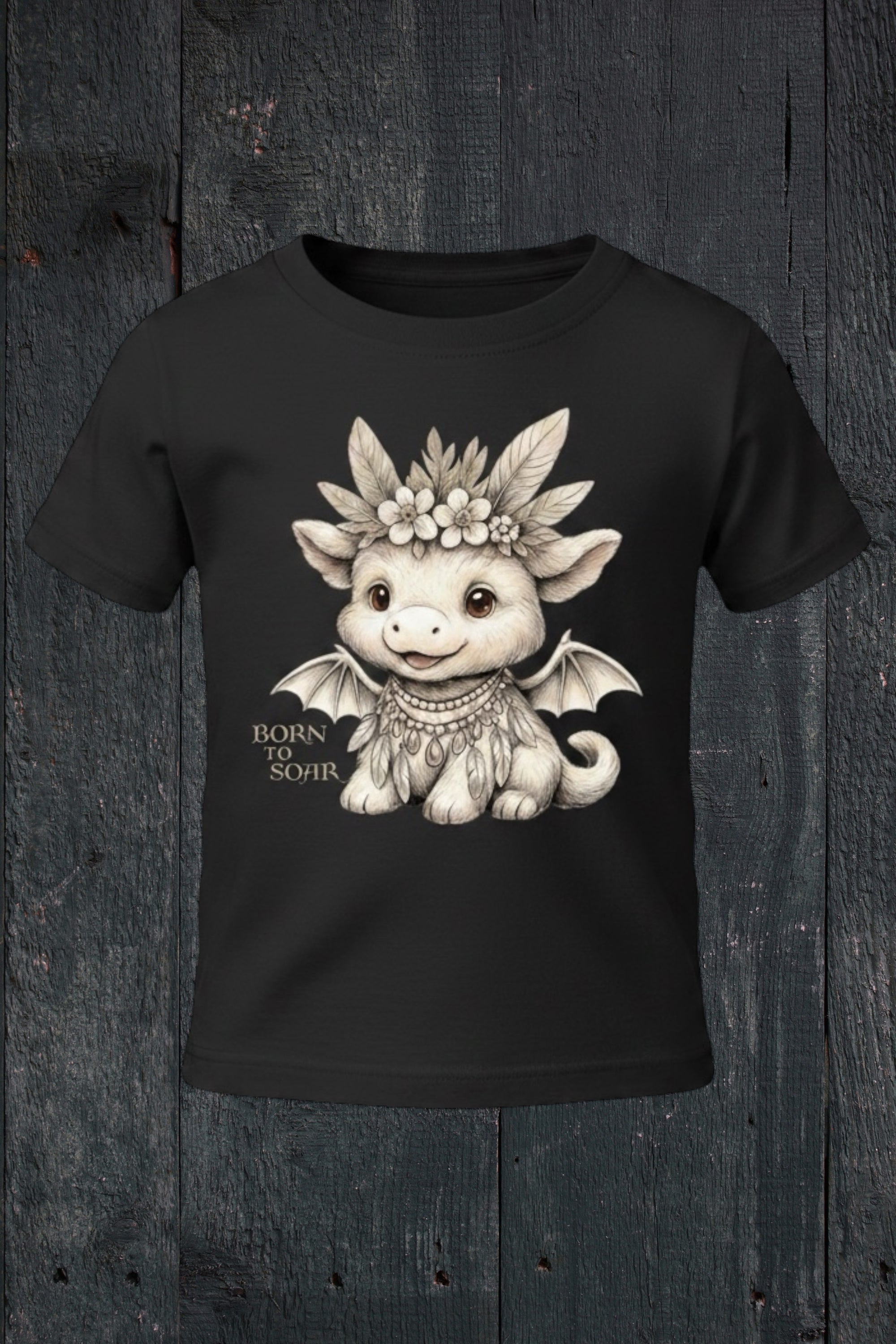 Flat lay of Fox and Form Ember the Mini Boho Dragon black classic ring spun tee with hand-illustrated boho dragon design and Born to Soar script on a dark wood background