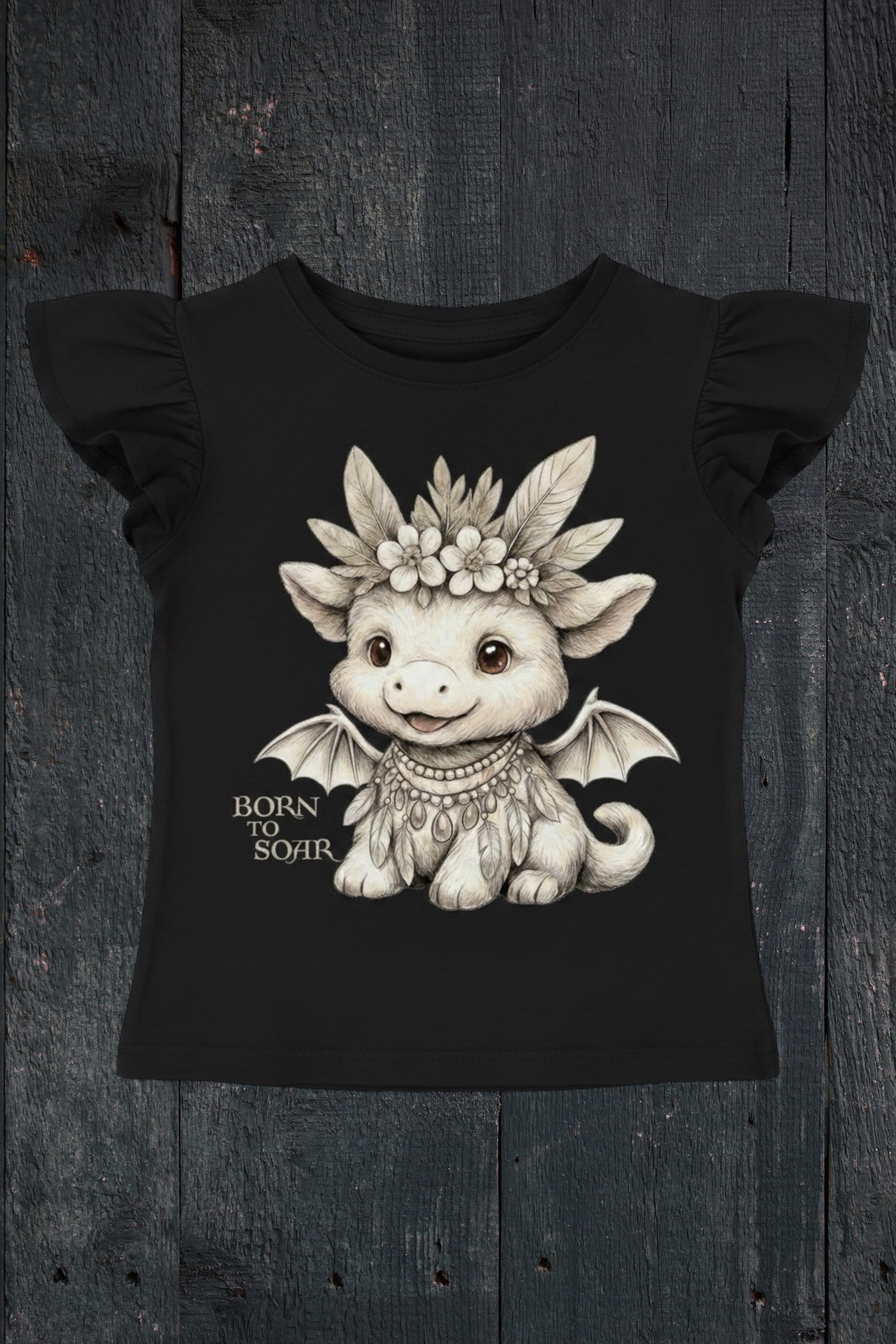 Flat lay of Fox and Form Ember the Mini Boho Dragon black flutter sleeve tee with hand-illustrated boho dragon design and Born to Soar script on a dark wood background