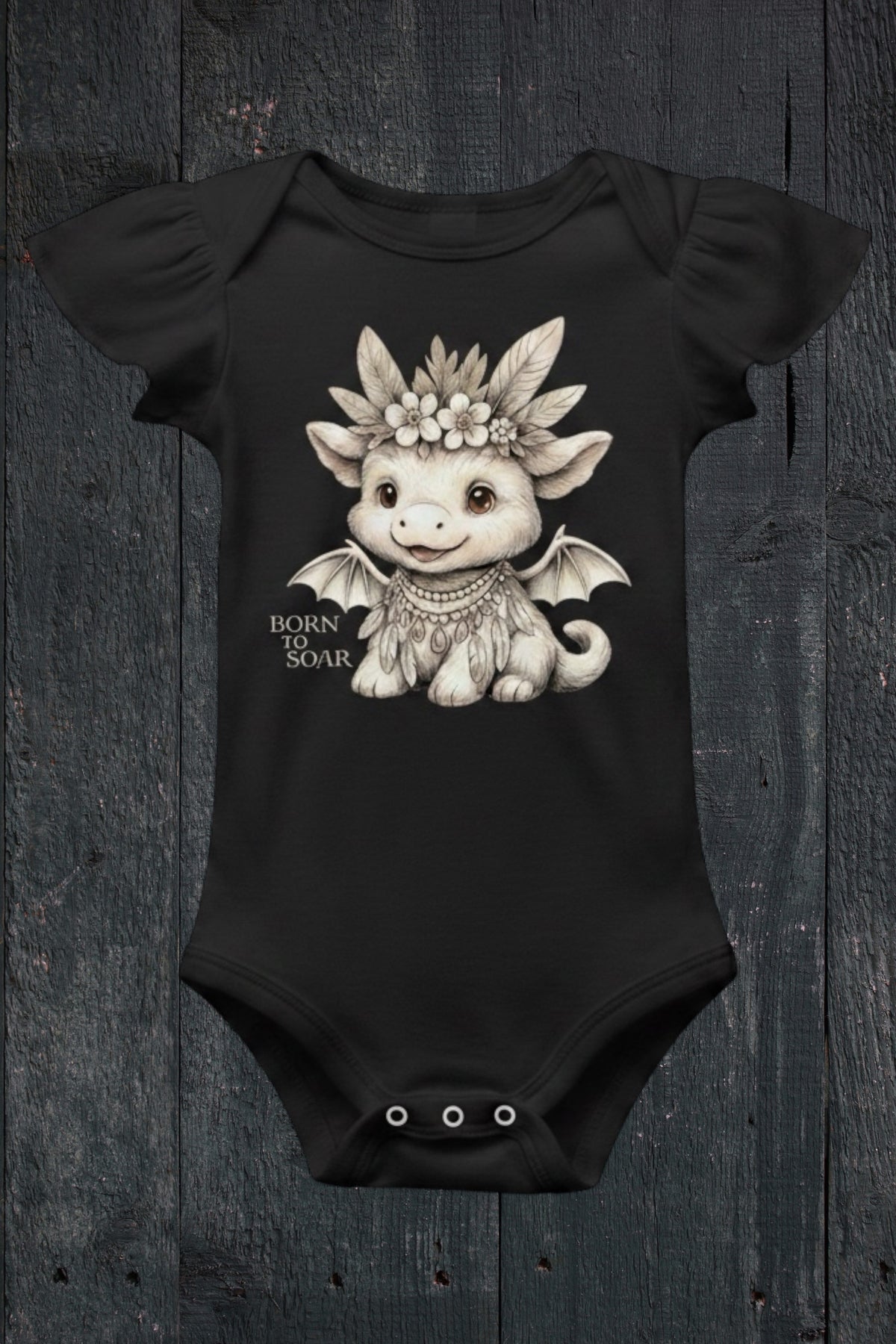 Flat lay of Fox and Form Ember the Mini Boho Dragon black flutter sleeve onesie with hand-illustrated boho dragon design and Born to Soar script on a dark wood background