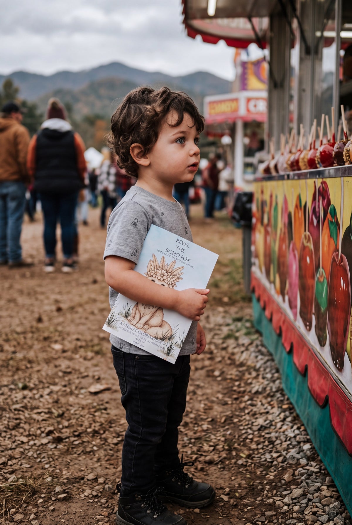 A young boy stands at a fair or carnival next to a candy stand, holding a book titled 'Revel the Boho Fox' in one hand and wearing a Fox & Form Sport Grey Fox hand-etched pattern classic tee shirt.  The background shows other fairgoers and mountains under an overcast sky. Fox & form. The Mini & youth Signature Collection. 