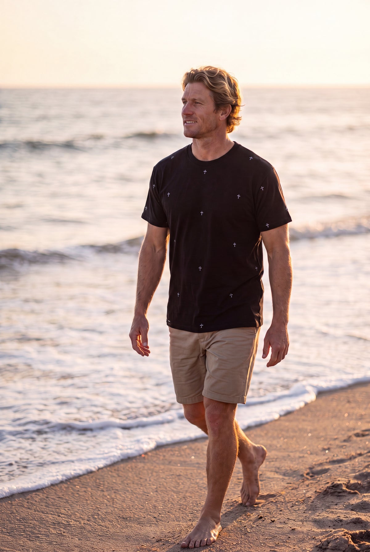 A man walks on a sandy beach at sunset wearing The Cross classic fit ring spun tee in black-hand illustrated cross pattern-Fox and Form. The Men's Signature Collection. 