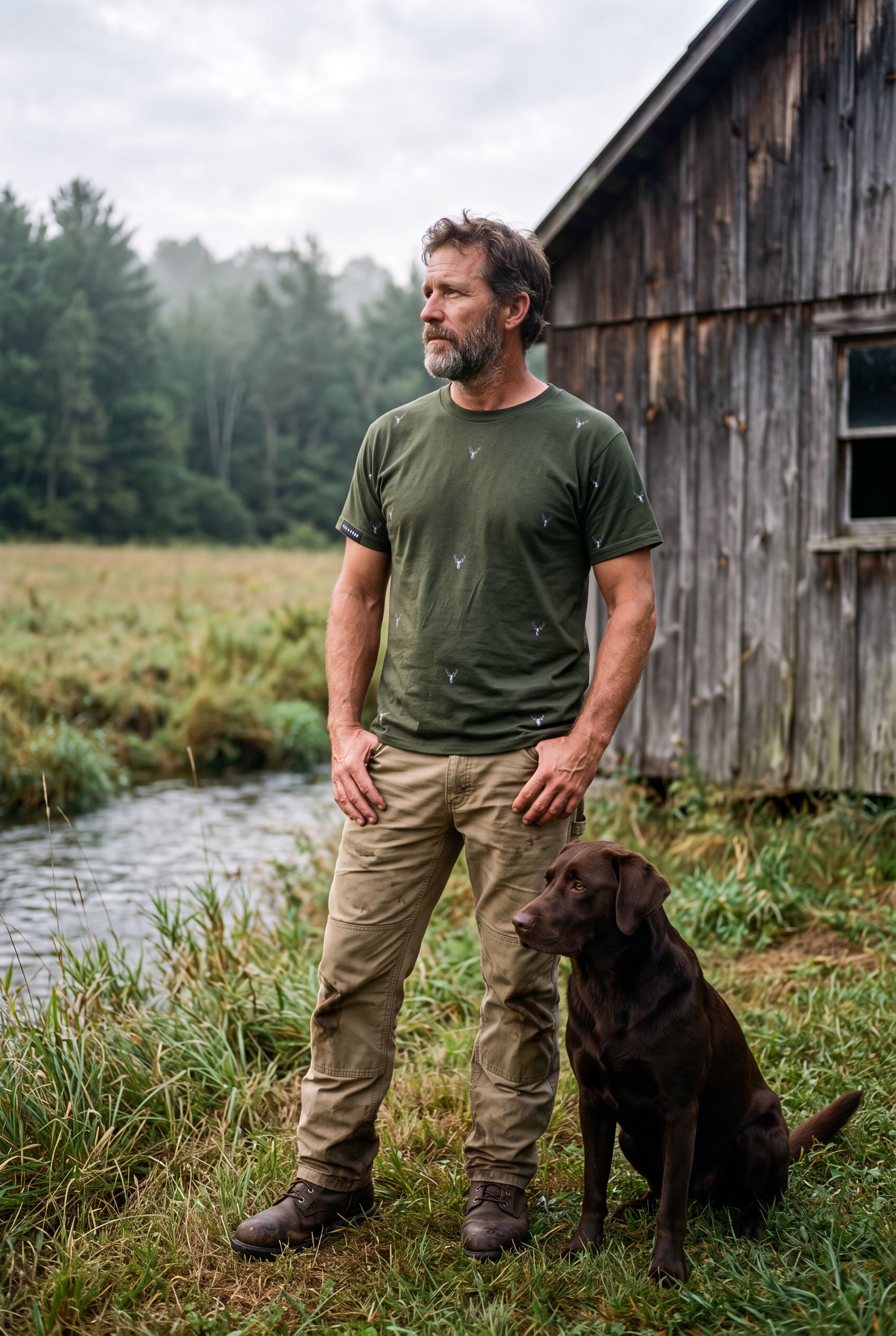 The Stag-men's hand illustrated forest green classic fit ring spun tee shirt worn outdoors next to a wooden cabin and a brown dog, with a stream and forest in the background. Fox & Form 
Men's Signature Collection
