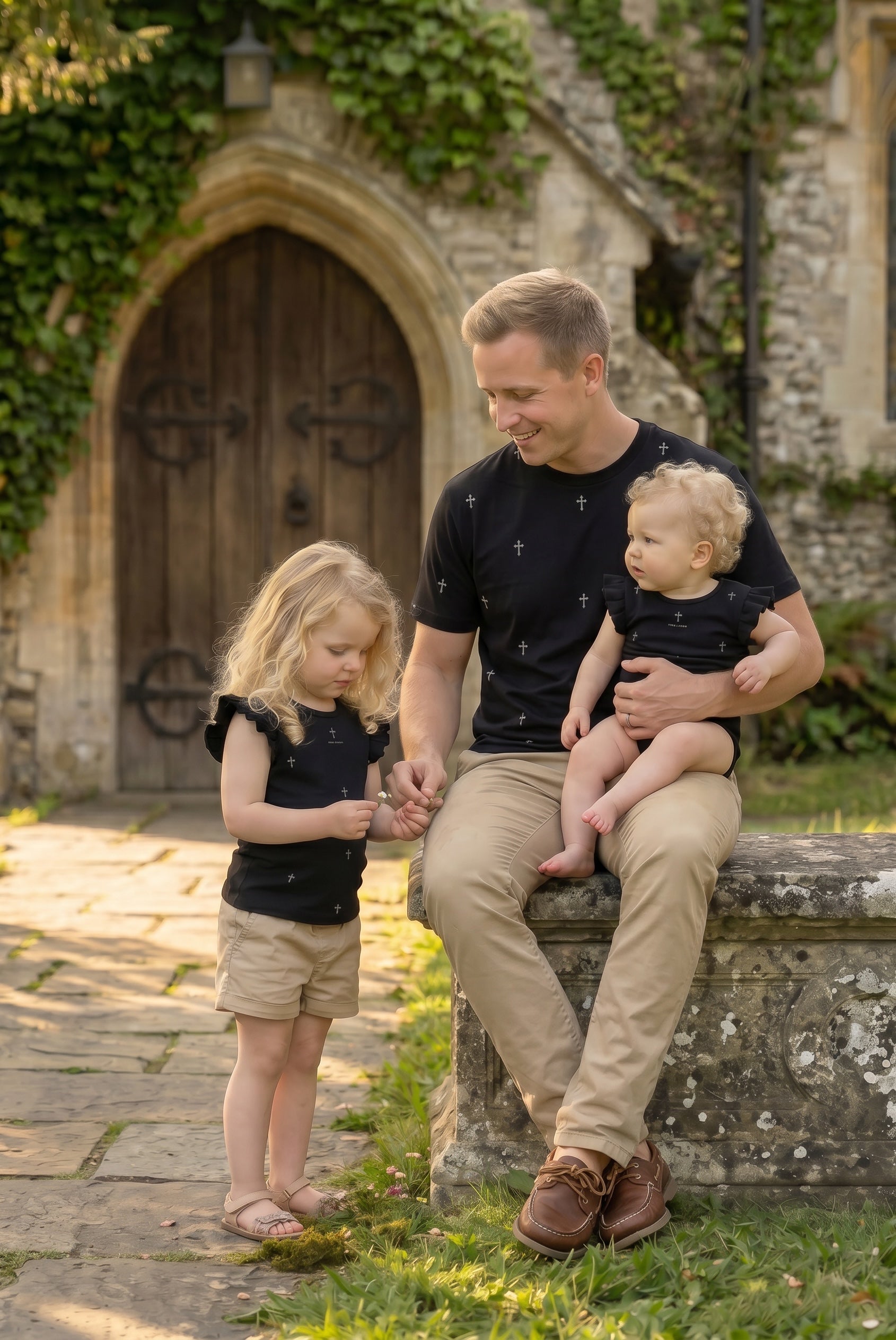 Man sitting on a stone bench with two children outdoors wearing classic fit ring spun tee shirt in black with hand illustrated cross pattern design and toddler girl wearing flutter sleeve tee shirt in black with hand illustrated cross pattern design and infant girl wearing flutter sleeve onesie in black with hand illustrated cross pattern design-Fox and  Form. Fox and Form Men's Signature Collection. Fox and Form Mini &amp; Youth Signature Collection. 