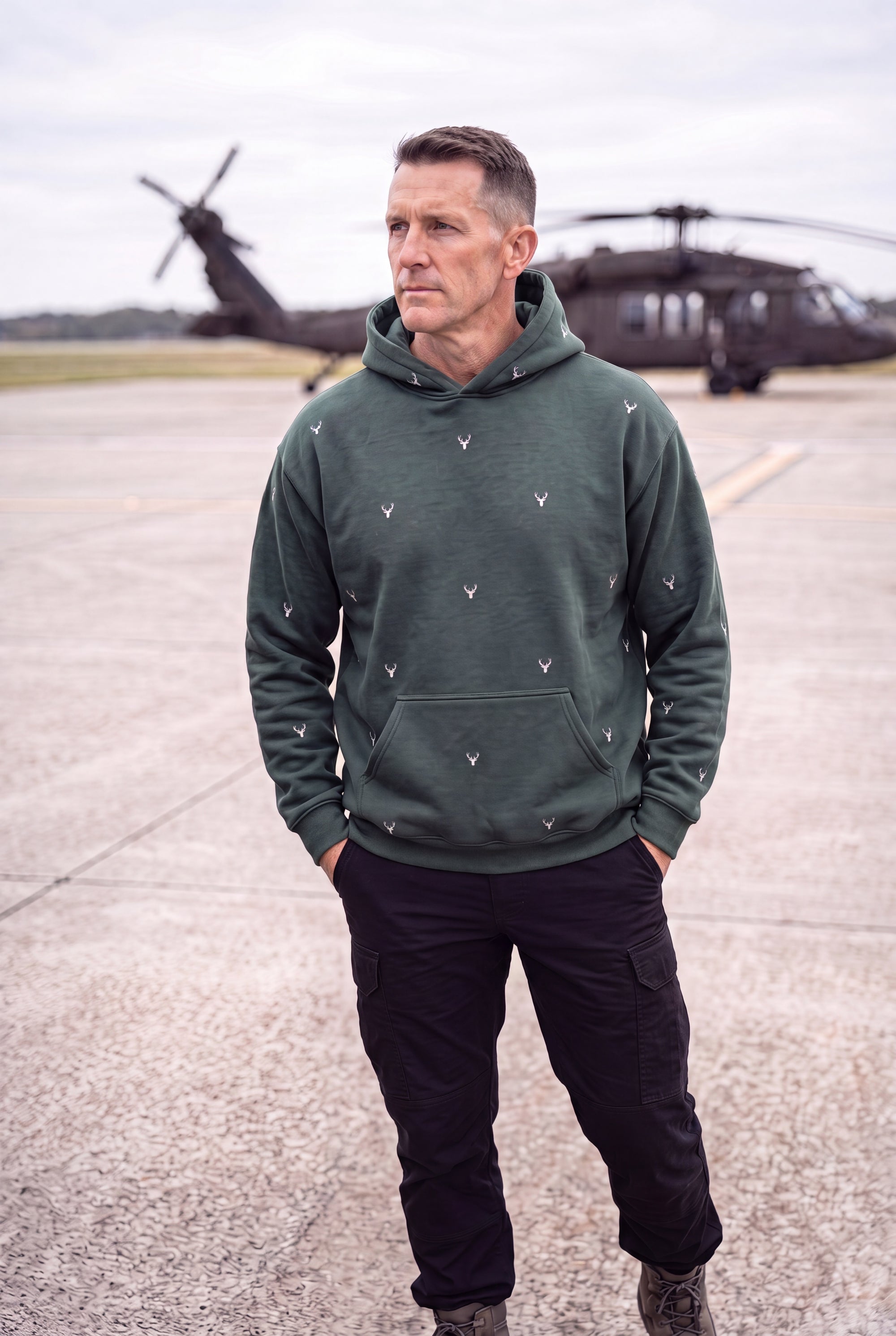 A man wearing Fox and Form The Stag Forest green dropped shoulder max weight sweatshirt with hood with hand-etched illustrated stag pattern design, standing with his hands in his pockets relaxed on an overcast day at a military helipad. Fox and Form Men's Signature Collection. 
