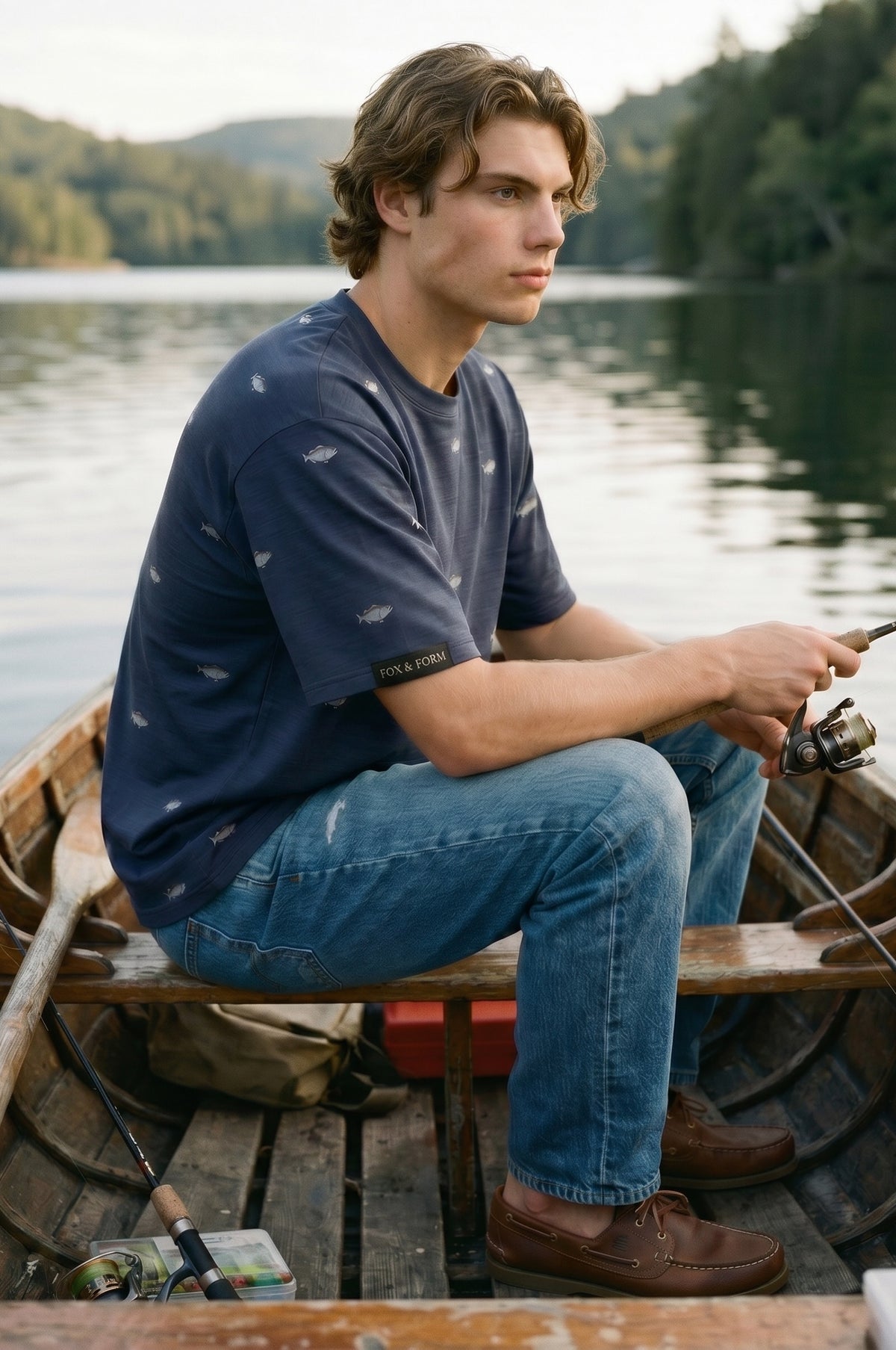 Young man wearing Fox and Form The Bass dropped shoulder max weight tee shirt in navy with hand-etched illustrated bass pattern design, holding a fishing rod in a wooden rowboat on a calm lake. Fox and Form Men's Signature Collection. 