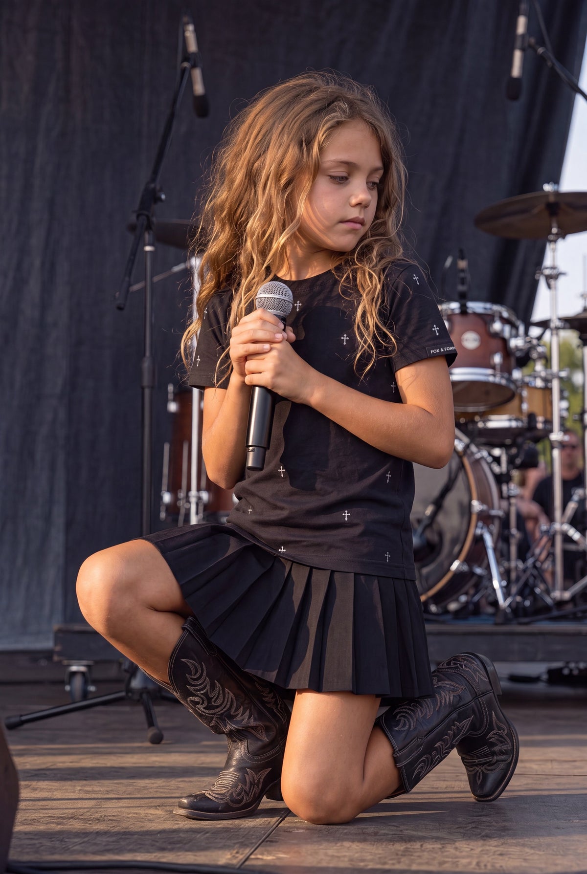 A young girl with long wavy light brown hair kneels on a stage holding a microphone. She is wearing a Fox and Form black classic fit ring spun short-sleeve t-shirt with hand-illustrated small cross details, a black pleated mini skirt, and black cowboy boots. Fox and Form. Mini & Youth Signature collection. 