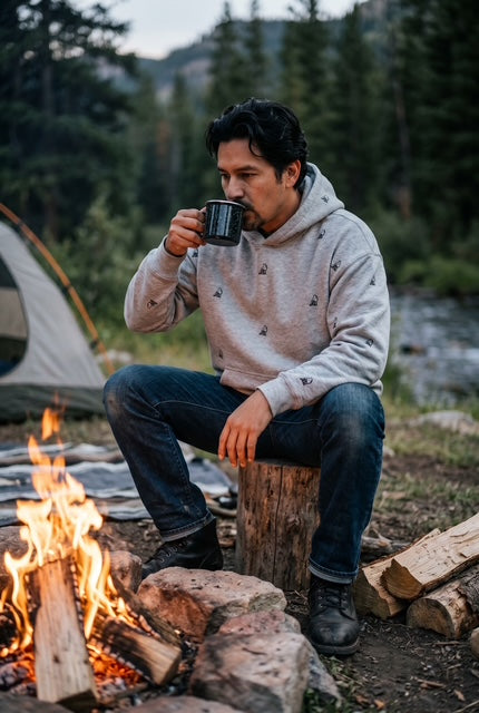 Man wearing The Fox sports grey max weight hooded sweatshirt with hand-etched illustrated fox pattern design, sitting by a campfire in a mountain forest at dusk. Fox and Form Men's signature Collection. 