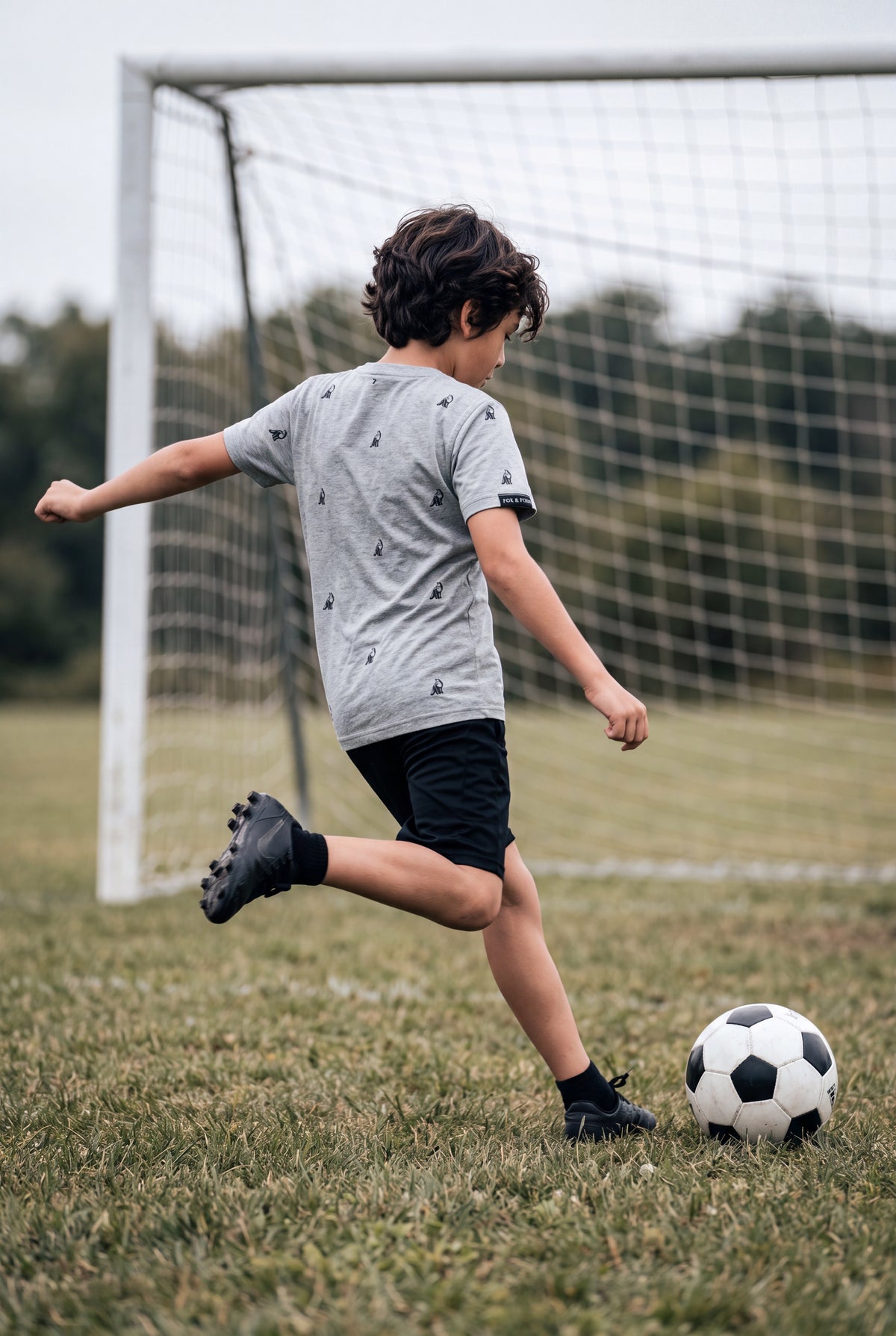 Fox and Form . A boy in a Fox and Form classic fit ring spun gray hand-illustrated fox patterned t-shirt and black shorts kicks a soccer ball on a grassy field in front of a soccer goal. Fox and Form. Mini & Youth Signature Collection. 