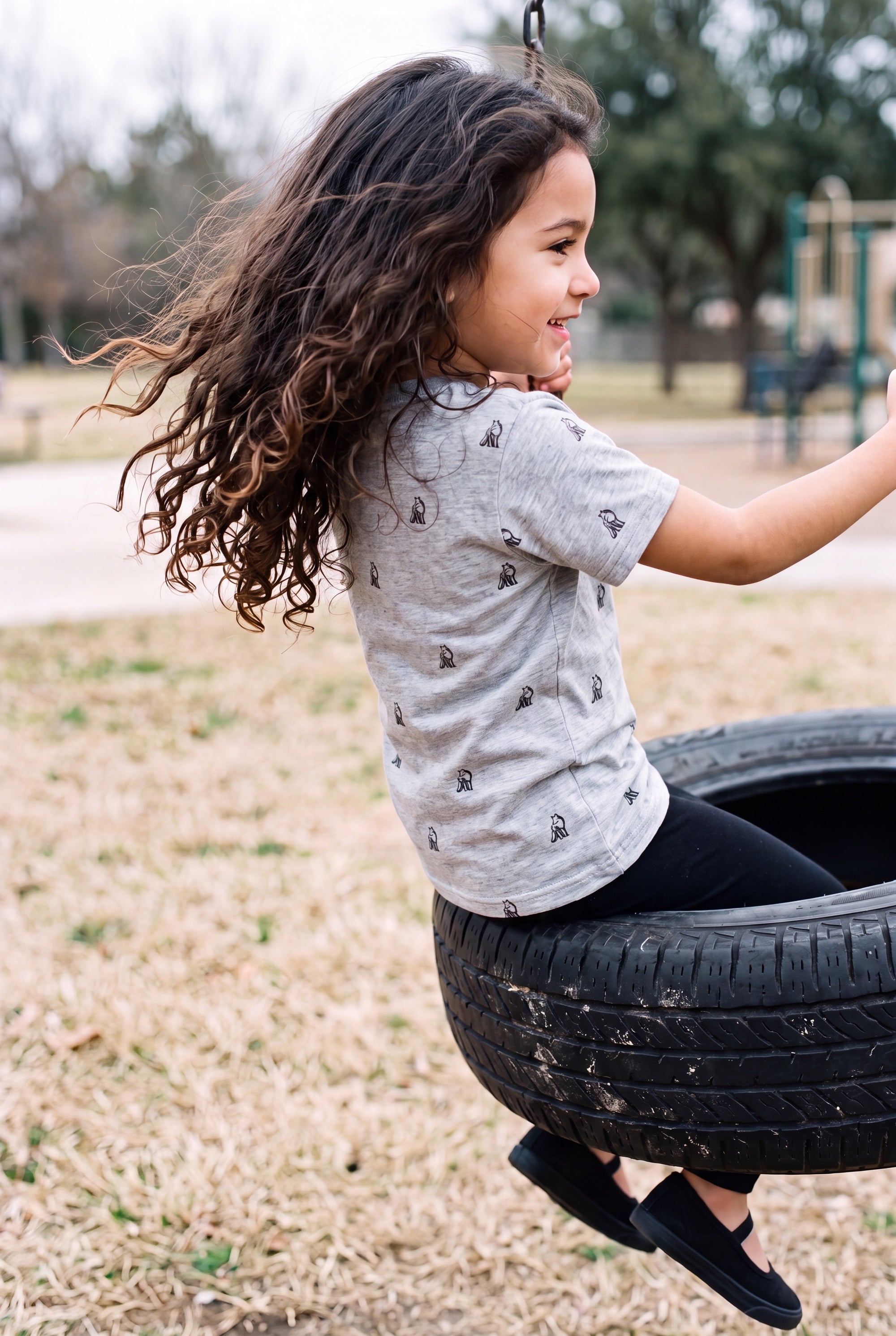 A young girl with long, curly brown hair sits on a tire swing at a playground, wearing a Fox and Form classic fit ring spun The fox sport gray short-sleeve t-shirt with small hand-illustrated foxes, black pants, and black slip-on shoes. Fox and Form. Mini & Youth Signature Collection. 