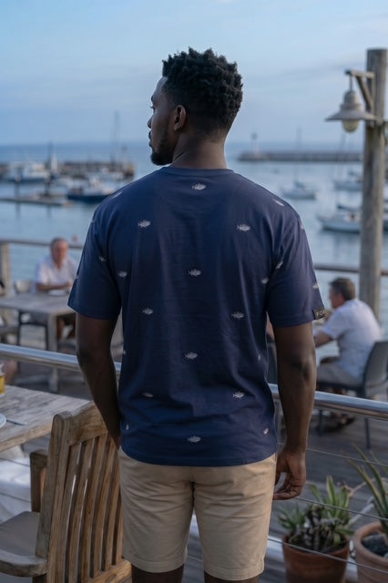 Back view of The Bass navy dropped shoulder max weight tee shirt with hand-etched illustrated bass pattern design, worn at a waterfront marina at dusk. Fox and Form Men's Signature Collection. 