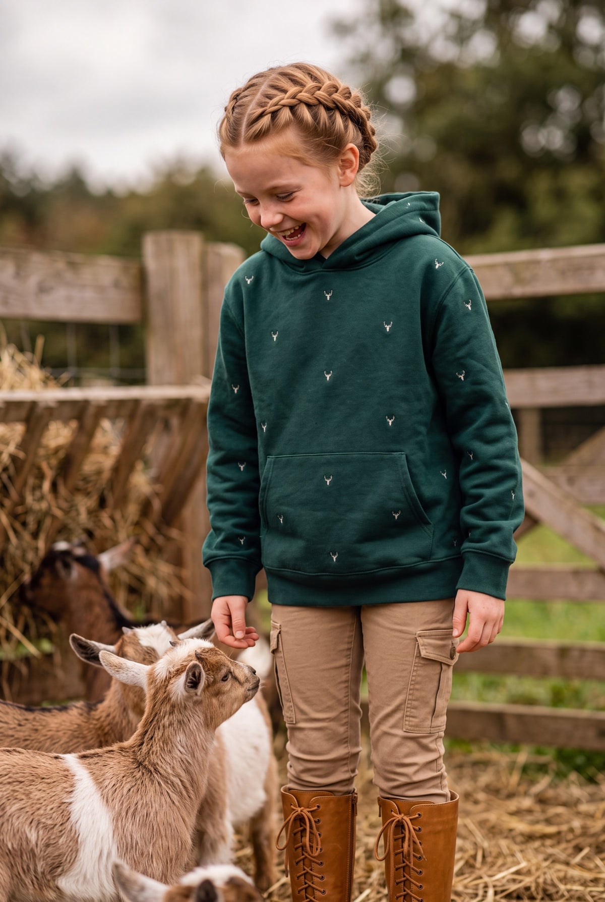A young girl with braided hair stands in a barn feeding a goat, wearing hand-etched Fox & Form Stag Forest green sweatshirt with small brown hand-etched stag pattern, tan cargo pants, and brown lace-up boots. Fox & Form. The Stag Youth Signature Collection.