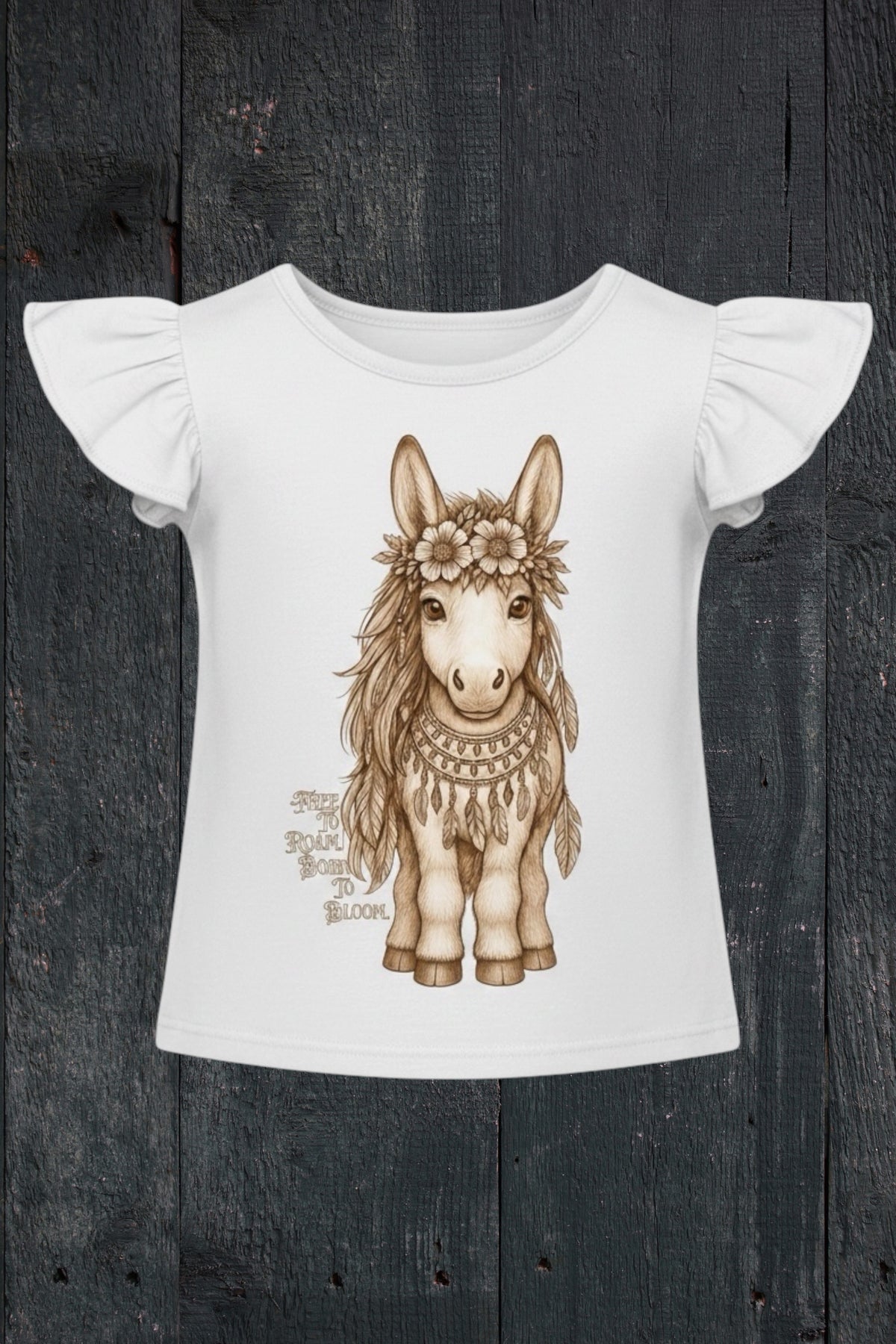 Flat lay of Fox and Form Meadow the Boho Pony white flutter sleeve tee with hand-illustrated boho pony design and Free to Roam Born to Bloom script on a dark wood background