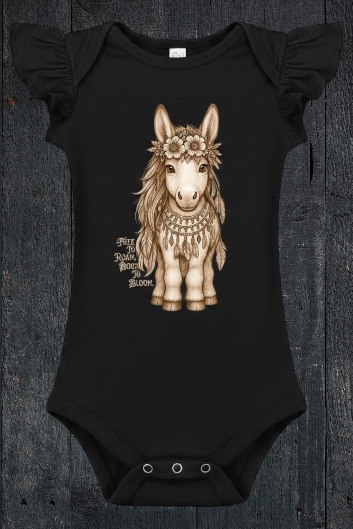 Flat lay of Fox and Form Meadow the Boho Pony black flutter sleeve onesie with hand-illustrated boho pony design and Free to Roam Born to Bloom script on a dark wood background