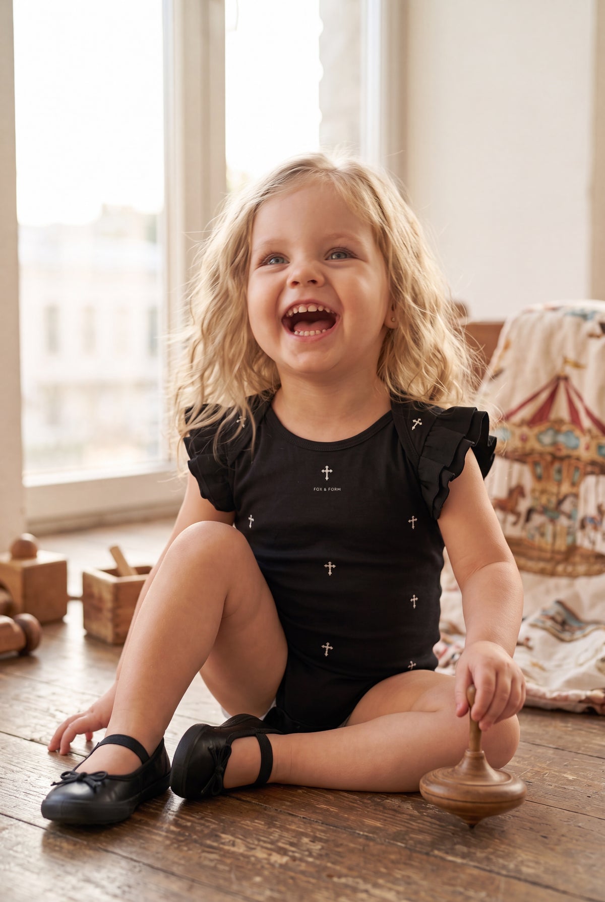 Toddler girl sitting on cobblestone path in front of a carousel wearing Fox and Form The Cross black flutter sleeve mini onesie with hand-etched illustrated cross pattern design