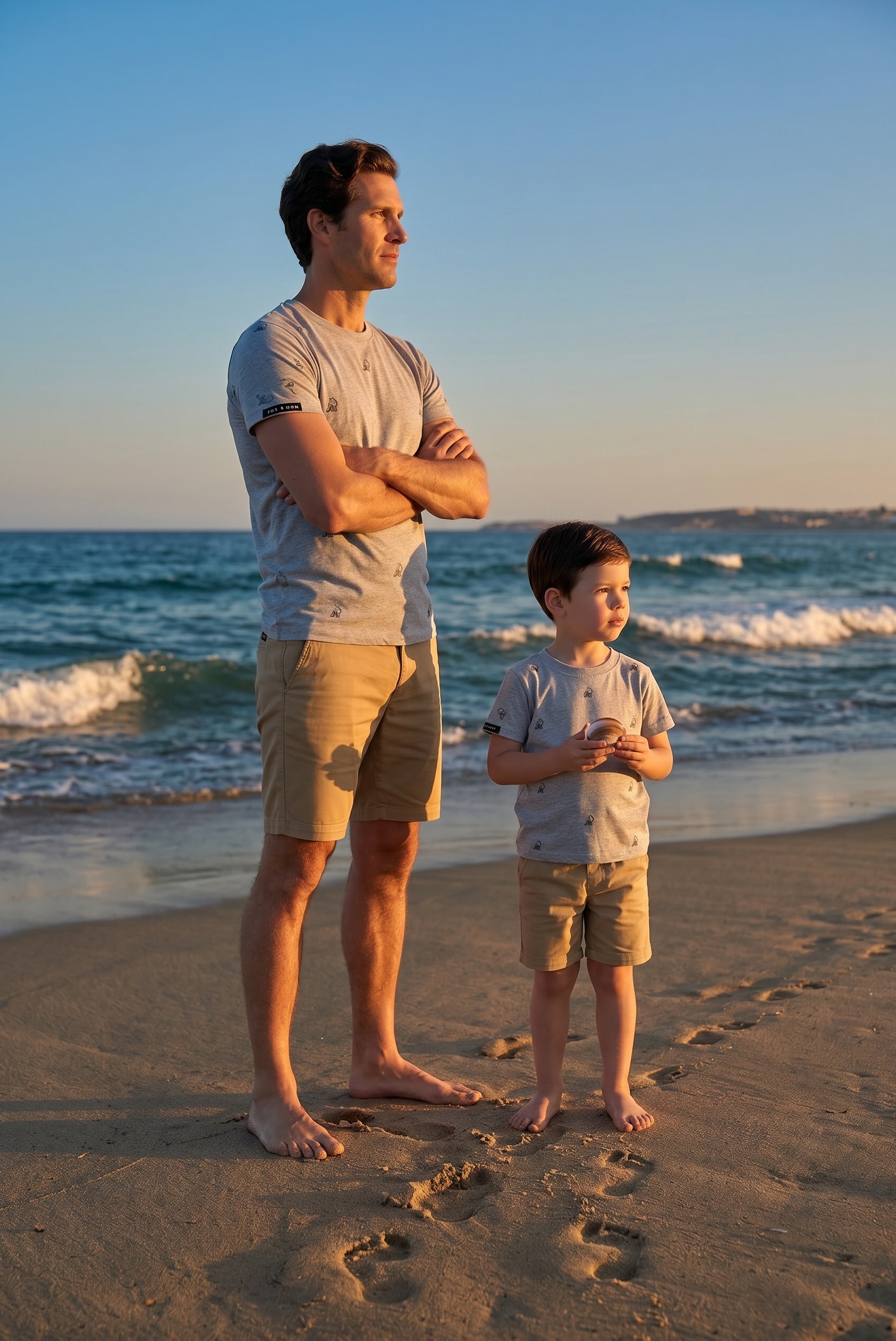 Father and son wearing matching Fox and Form The Fox sports grey classic ring spun tee with hand-etched illustrated fox pattern design, standing together on a sandy beach at golden hour. Fox and Form Men's Signature Collection. Mini & Youth Signature Collection. 