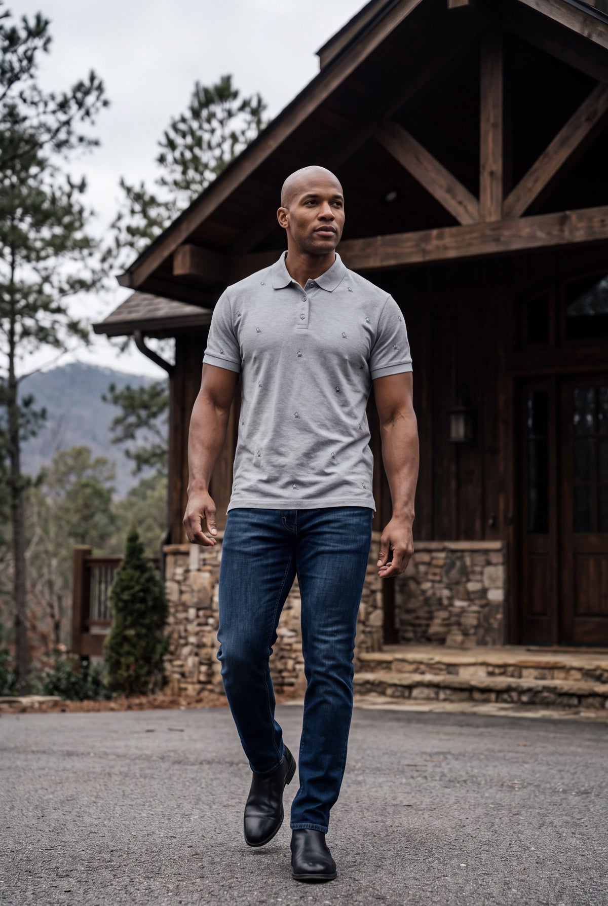 A man walking outdoors in front of a rustic wooden cabin, wearing a light gray short-sleeve polo shirt with hand-etched small foxes patterned on the front and back, dark blue jeans, and black boots. Fox & Form 