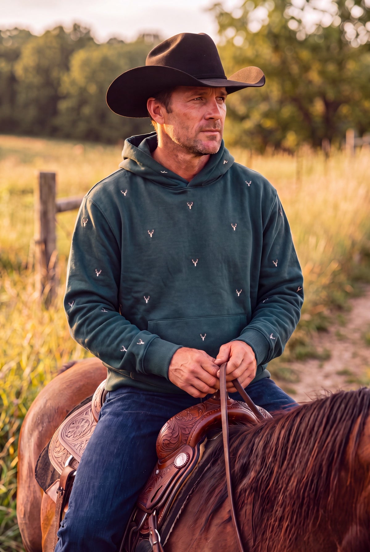 A man wearing a forest green double layered hoodie with a small brown hand-etched deer head patterned on the front, back and hood and a black cowboy hat, sitting on a brown horse in an outdoor field at sunset. Fox & Form 