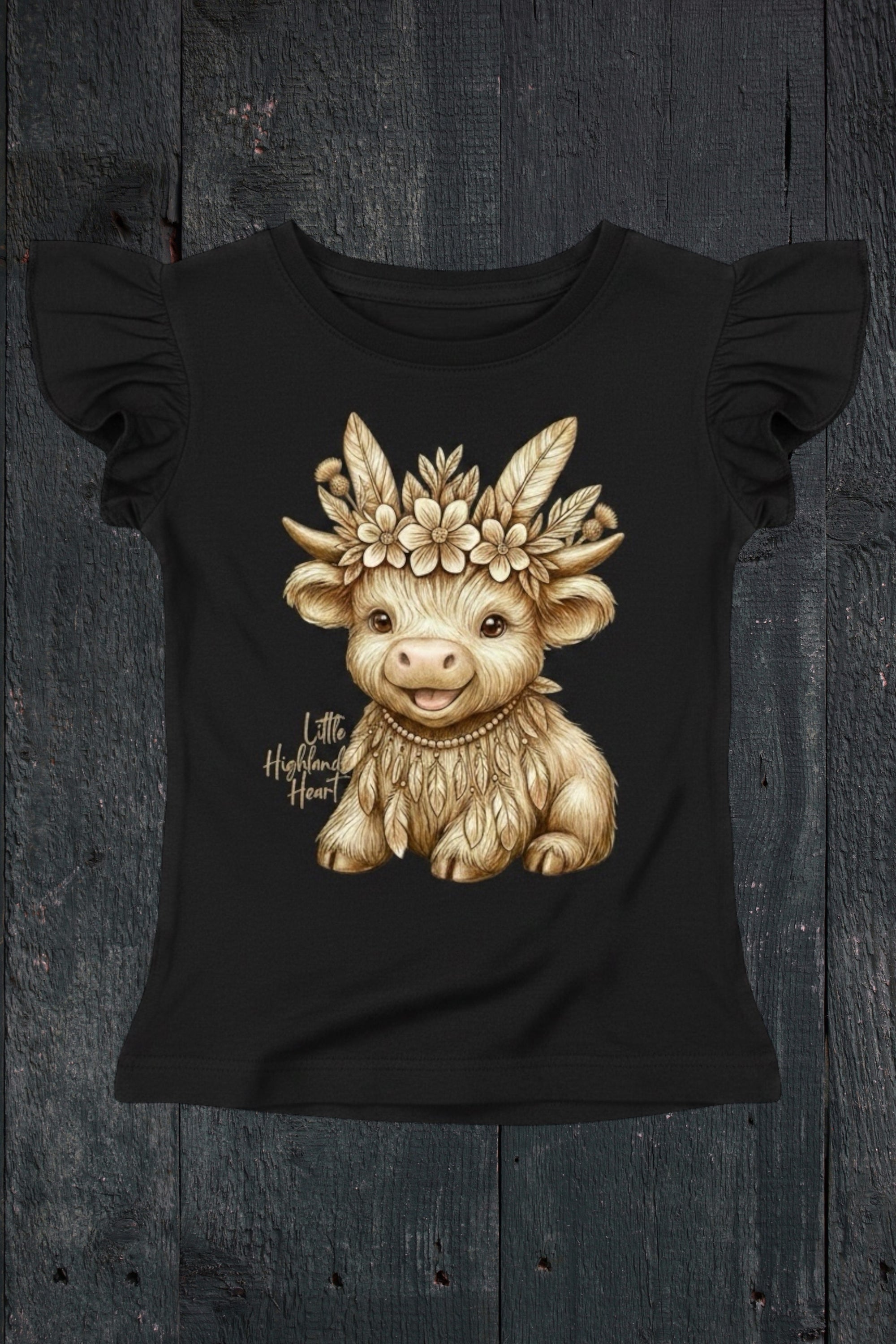 Flat lay of Fox and Form Thistle the Boho Highland Calf black flutter sleeve tee with hand-illustrated boho highland calf design and Little Highland Heart script on a dark wood background