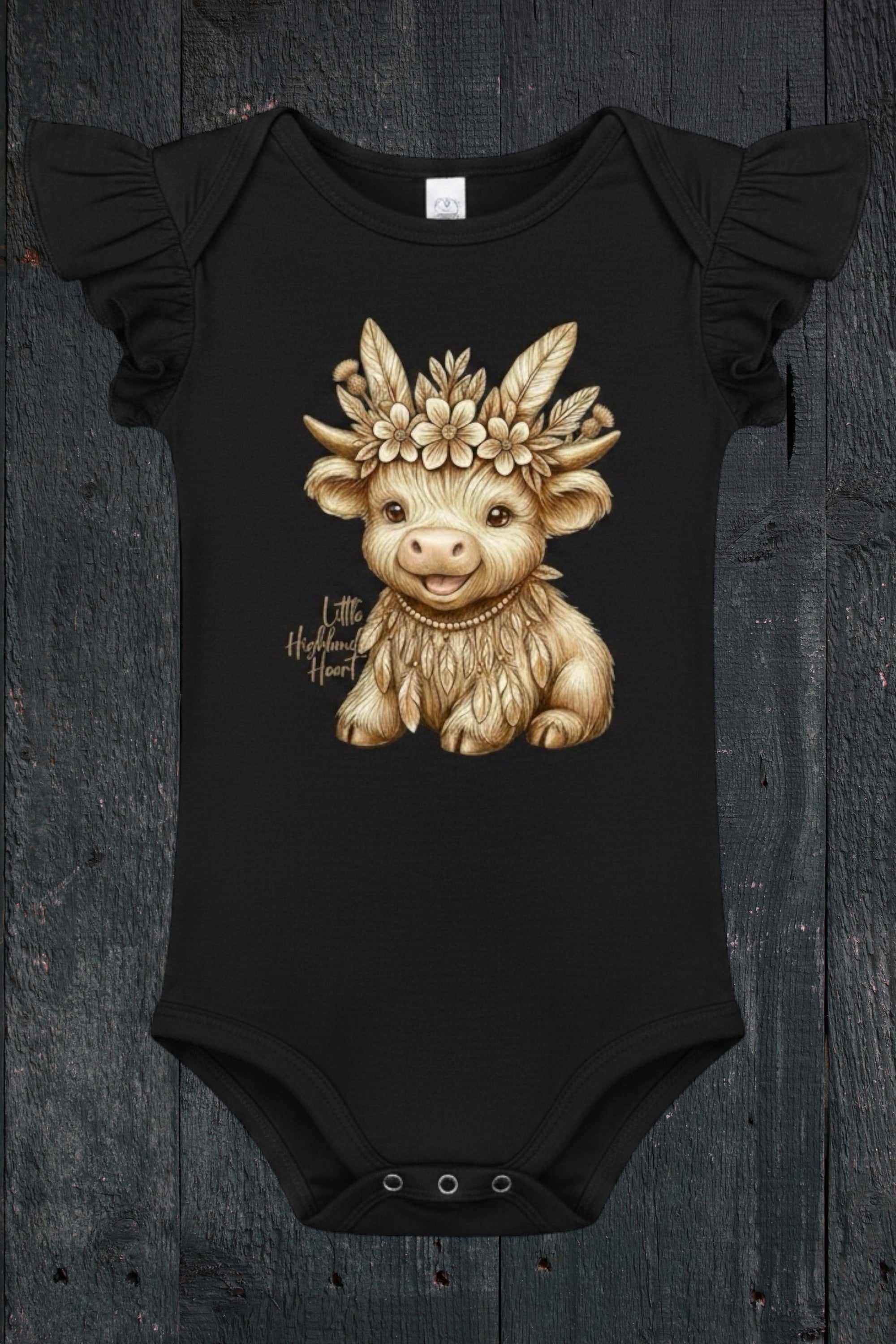Flat lay of Fox and Form Thistle the Boho Highland Calf black flutter sleeve onesie with hand-illustrated boho highland calf design and Little Highland Heart script on a dark wood background