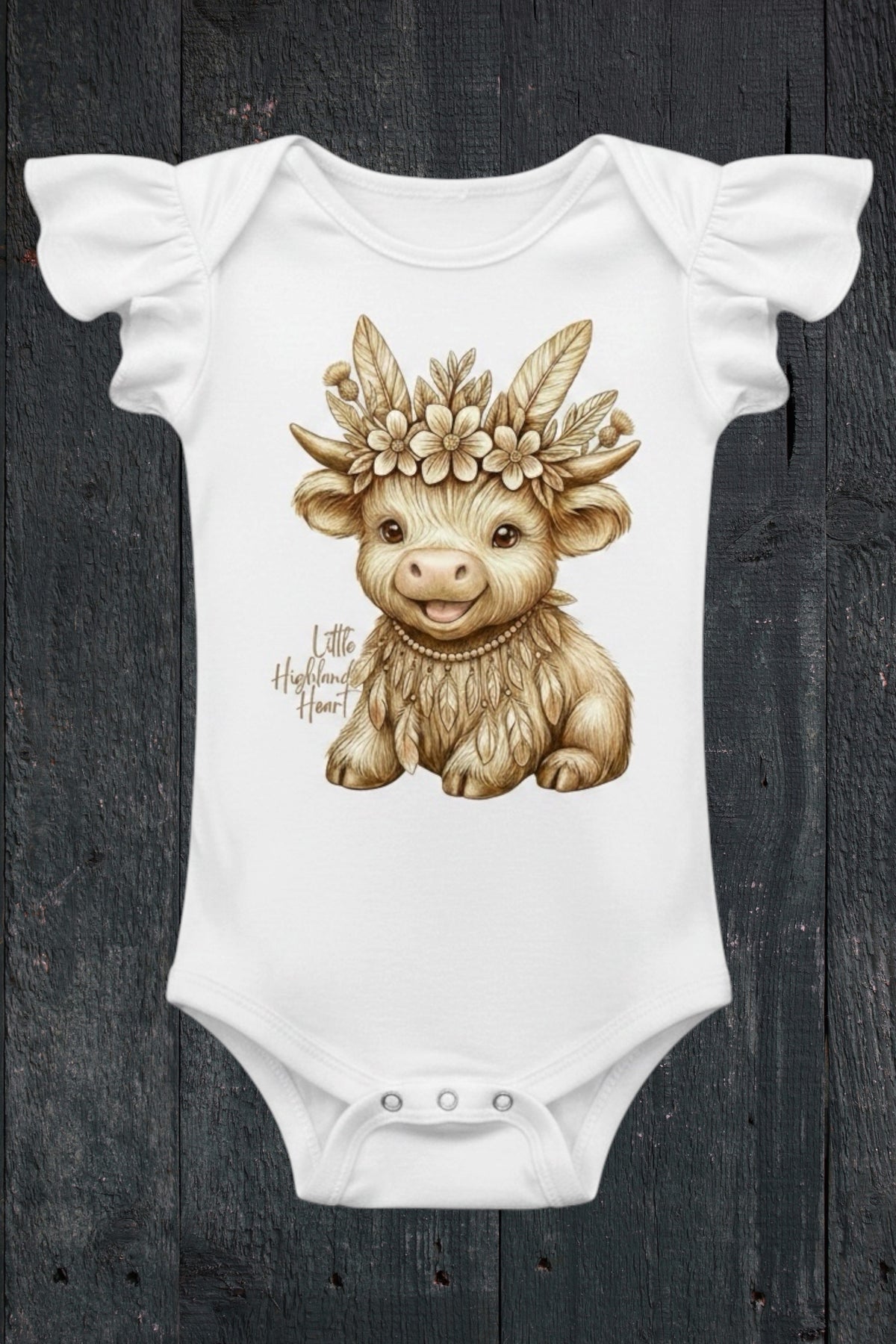 Flat lay of Fox and Form Thistle the Boho Highland Calf white flutter sleeve onesie with hand-illustrated boho highland calf design and Little Highland Heart script on a dark wood background