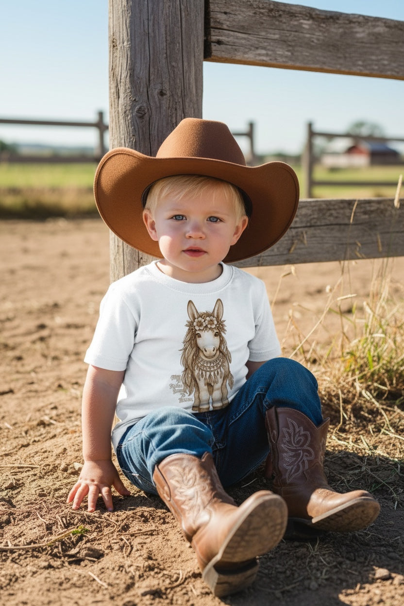 Toddler boy wearing a brown cowboy hat and cowboy boots sitting against a wooden ranch fence post on a sunny day wearing Fox and Form Meadow the Boho Pony white classic ring spun tee with hand-illustrated boho pony design and Free to Roam Born to Bloom script